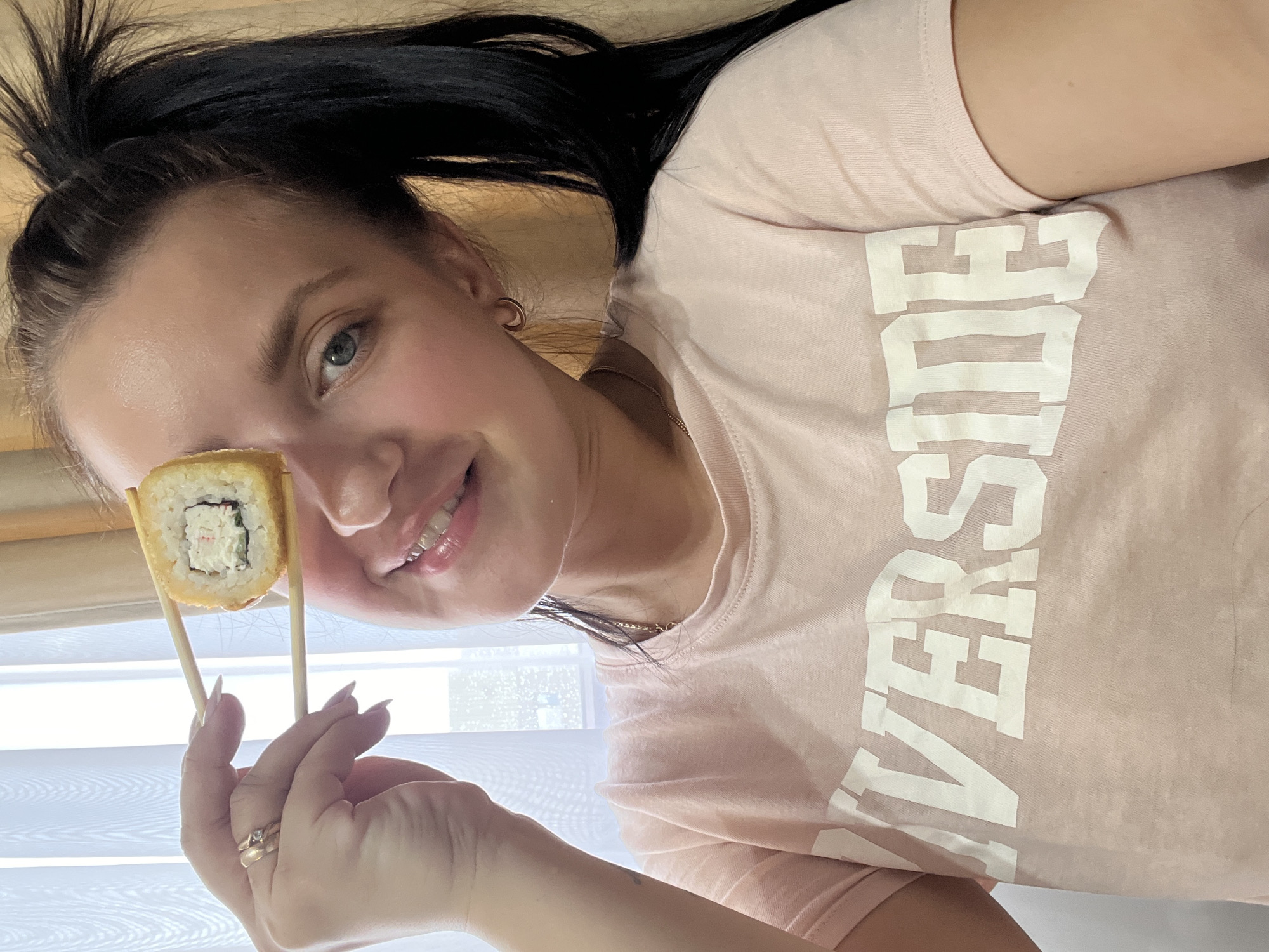 A young woman with dark hair pulled into a ponytail and blue eyes is holding a piece of sushi up to her face with chopsticks, partially obscuring her left eye. She is wearing a light pink t-shirt with the word "RIVERSIDE" printed in white block letters. She is smiling, and her makeup is visible. The background appears to be indoors, with sheer white curtains on the left and a muted tan wall or curtain on the right. The lighting suggests it is daytime, possibly near a window. The location is not specified within the image itself.