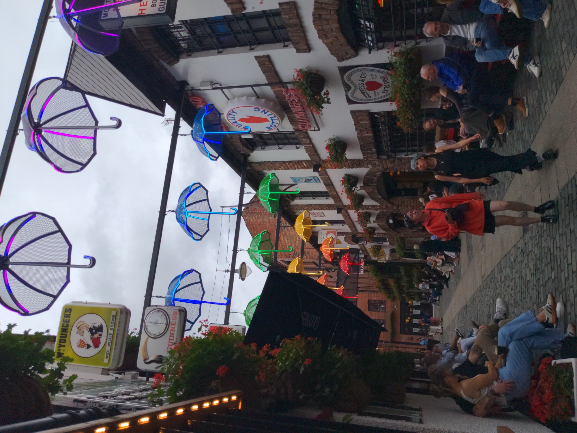 The image depicts a street scene, likely a pedestrian alley or courtyard, with numerous people engaged in various activities. Overhead, a series of decorative umbrellas are suspended, many of which are illuminated with colorful LED lights, including blue, green, purple, and pink. The architecture suggests a historic European setting, with brick buildings lining the sides of the alley. Several signs are visible, including a yellow sign for "WEE WILLIE BROWN" and a Guinness clock.
Several individuals are seated on a low wall or bench along the left side of the alley, some in conversation, others looking out. On the right side, a group of people are also seated, and some are standing. The ground is paved with cobblestones.
The scene appears to be a lively public space, possibly a street in the Cathedral Quarter of Belfast, Northern Ireland, known for its pubs, restaurants, and vibrant atmosphere. The presence of Guinness branding strongly suggests a connection to the famous Irish stout. The overall impression is one of a bustling, social outdoor area, decorated with artistic lighting.