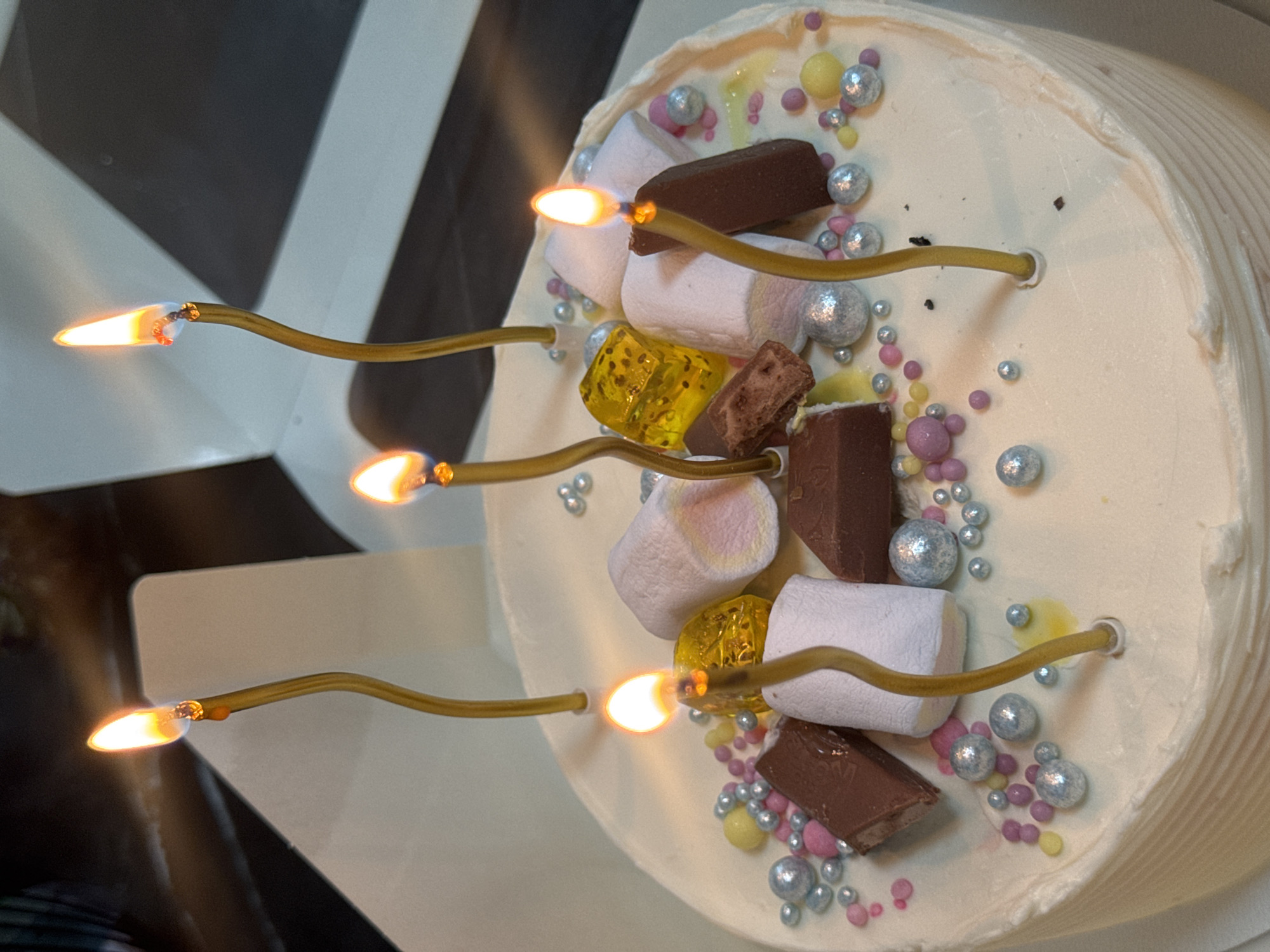 A close-up, eye-level shot shows a white cake adorned with an assortment of toppings. Scattered across the frosting are small, colorful candies in shades of pink, yellow, and silver, along with chunks of chocolate and marshmallows. Five golden candles, each lit with a flickering flame, are positioned upright on the cake. One candle is positioned in the center foreground, with three others arranged around it, and one more further back to the left. The background is out of focus, showing what appears to be a black and white striped surface, possibly a box lid. The lighting suggests an indoor setting, with the candles providing the primary light source. There are no people visible in the image. The image does not contain any visual elements that would allow for the determination of its location within Chaikovsky, Russia.