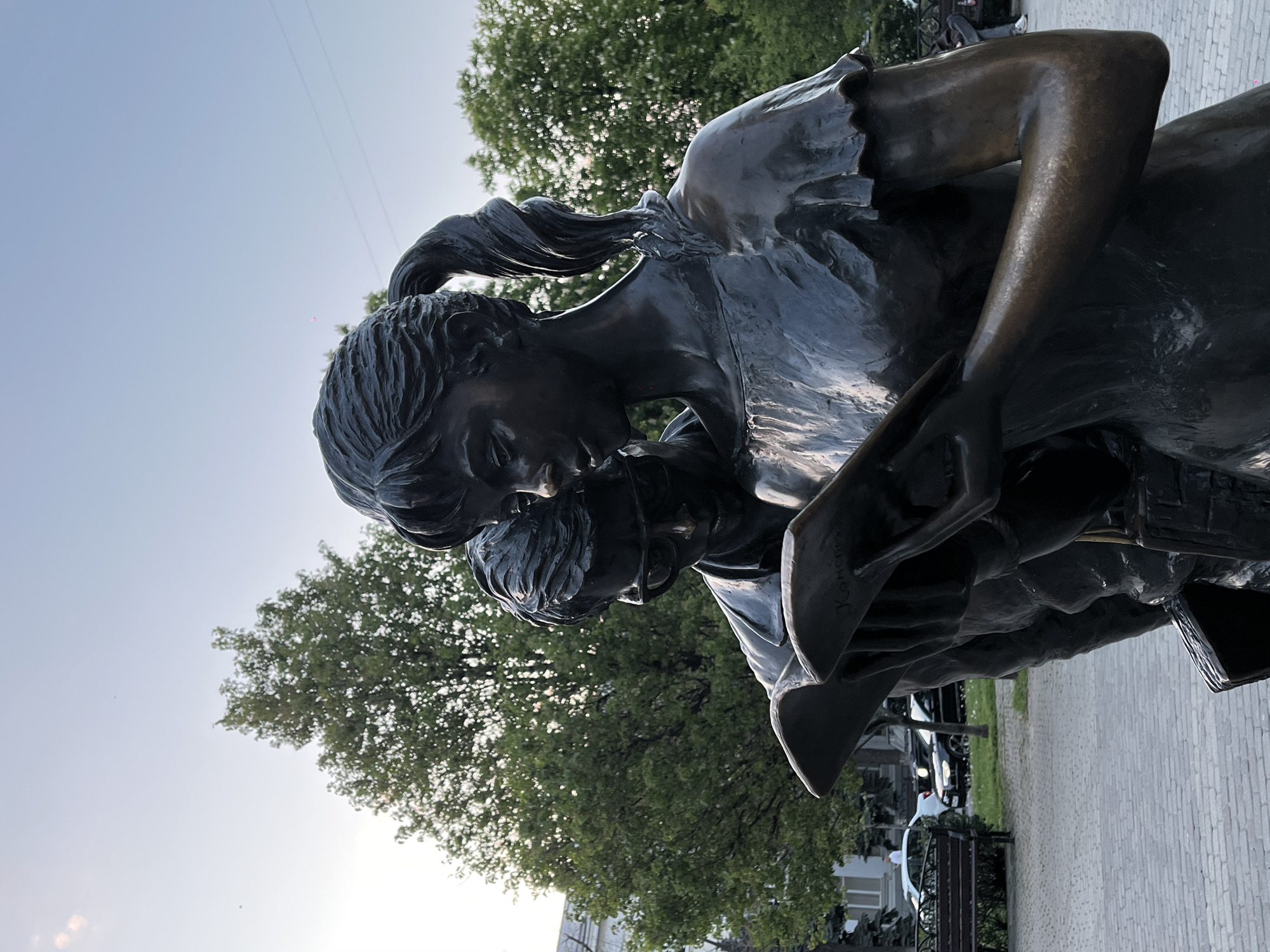 A bronze statue depicting two figures is the central subject, viewed from a low angle against a clear sky. The larger figure, likely an adult woman with hair pulled back and flowing locks, is shown in a tender embrace, holding a smaller figure, presumably a child, close to her chest. The woman looks downwards, and her hands hold an object resembling a book or tablet.
The activity portrayed is one of intimate connection, suggesting comfort, storytelling, or nurturing. The child's head is nestled into the woman's shoulder, conveying a sense of security.
Notable details include the statue's dark, reflective bronze material, which highlights the sculpted details of hair, clothing, and facial features. The background features lush green trees and a pale blue sky, indicating clear weather. Below the statue, a paved walkway or plaza is visible, with parked cars and distant buildings suggesting an urban park or public square setting. The lighting suggests late afternoon or early evening. The overall mood is one of peace and affection.
Visible text can be found on the edge or spine of the object held by the woman. The Cyrillic inscription "Коновал" (Konoval) is clearly legible, likely representing the sculptor's signature or a dedication.