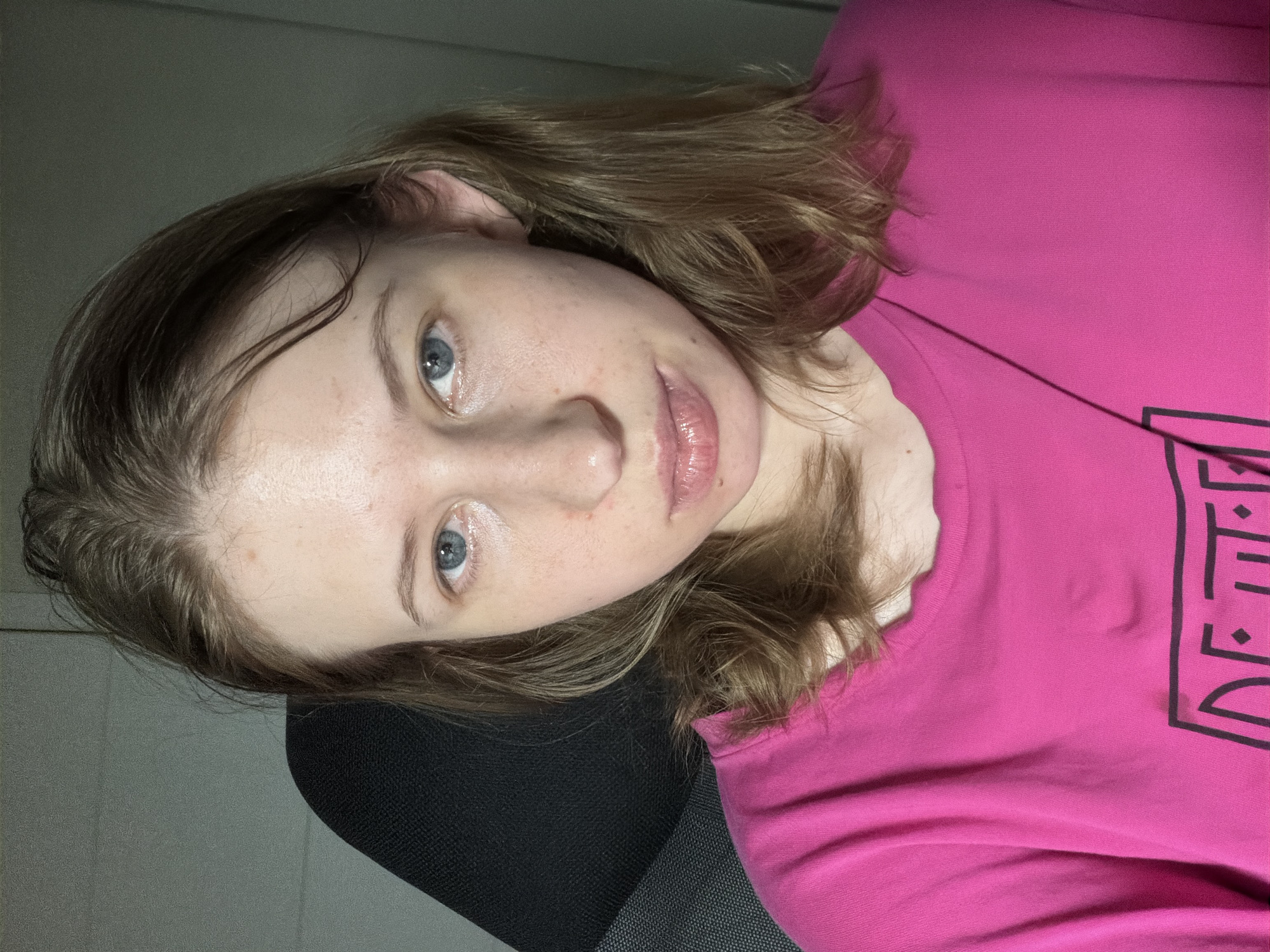 A young woman with shoulder-length blonde hair and blue eyes looks directly at the camera. She is wearing a bright pink t-shirt with black lettering on the front. The woman is seated in what appears to be an office chair with a black padded back. The background consists of a plain, grey wall, suggesting an indoor setting, likely a home or office environment in Dongducheon-si, South Korea. The lighting is direct, creating some shadows on the wall behind her and highlighting the texture of her skin.