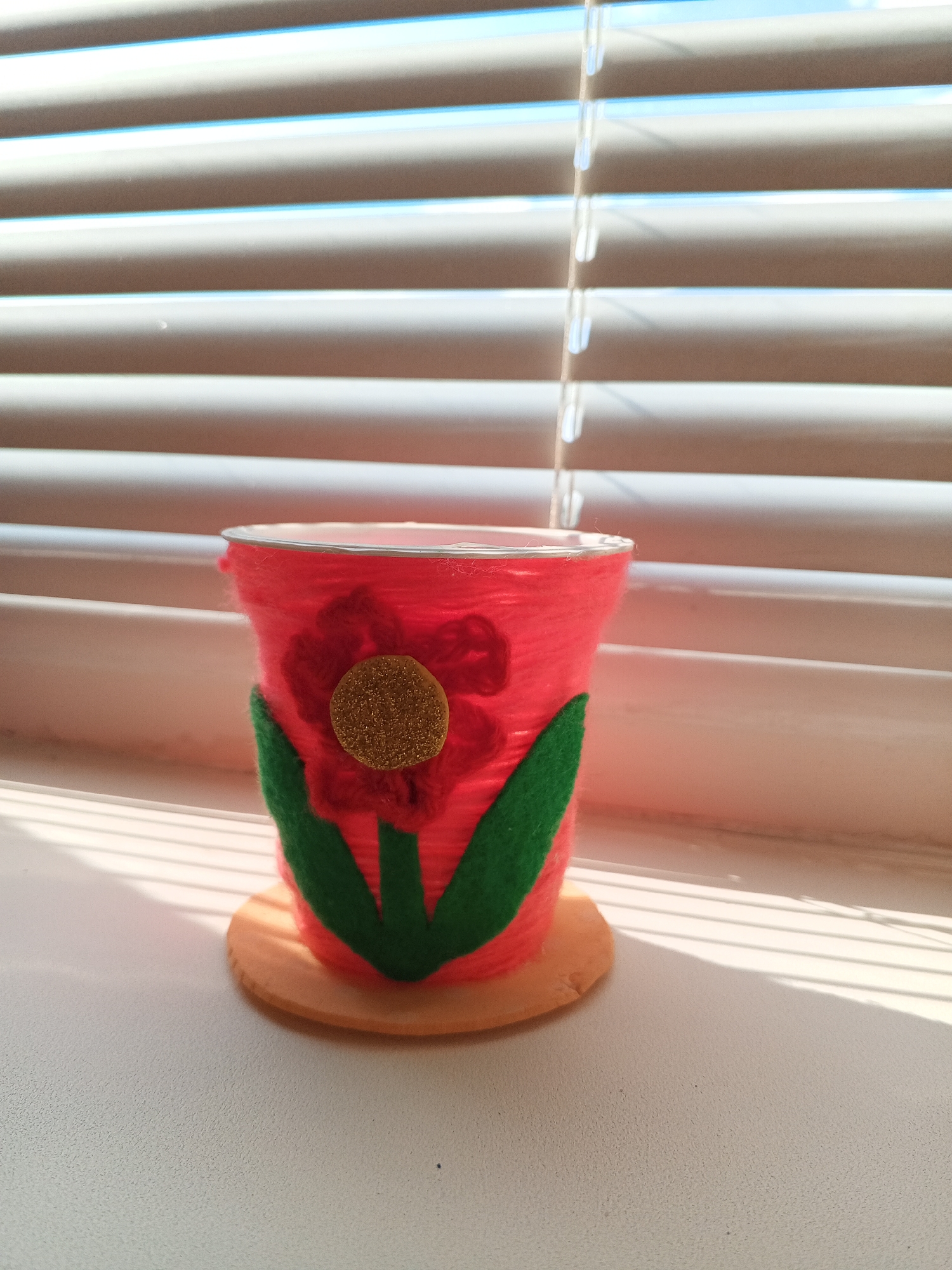 A close-up, eye-level shot shows a red, yarn-wrapped cup decorated with a felt flower, resting on a light brown circular base. The cup is adorned with a red flower with a gold glitter center and green felt leaves. The cup sits on a white surface in front of a set of closed beige blinds. Sunlight streams through the blinds, casting striped shadows across the scene. The background consists of the blinds and a sliver of blue sky. The image is set in Chistopol, Russia.