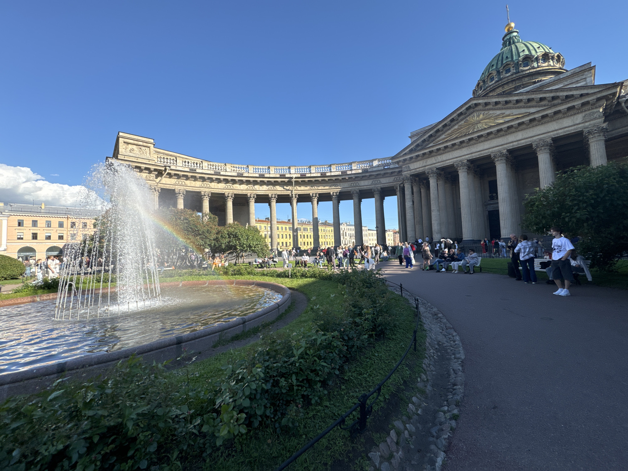 The image depicts a sunny day scene at Kazan Cathedral in Saint Petersburg, Russia. The grand neoclassical cathedral, with its iconic semicircular colonnade and green dome topped with a golden spire, dominates the background. In the foreground, a fountain sprays water, creating a rainbow. People are scattered throughout the scene, some walking along the paved pathway, others sitting on benches or standing in small groups. The atmosphere appears relaxed and pleasant, suggesting a typical tourist activity or a leisurely outing. The time of day is likely midday or early afternoon, given the bright sunlight and clear blue sky. The presence of lush green bushes and trees adds to the picturesque setting. A building with a light-colored facade and multiple windows is visible behind the colonnade.
