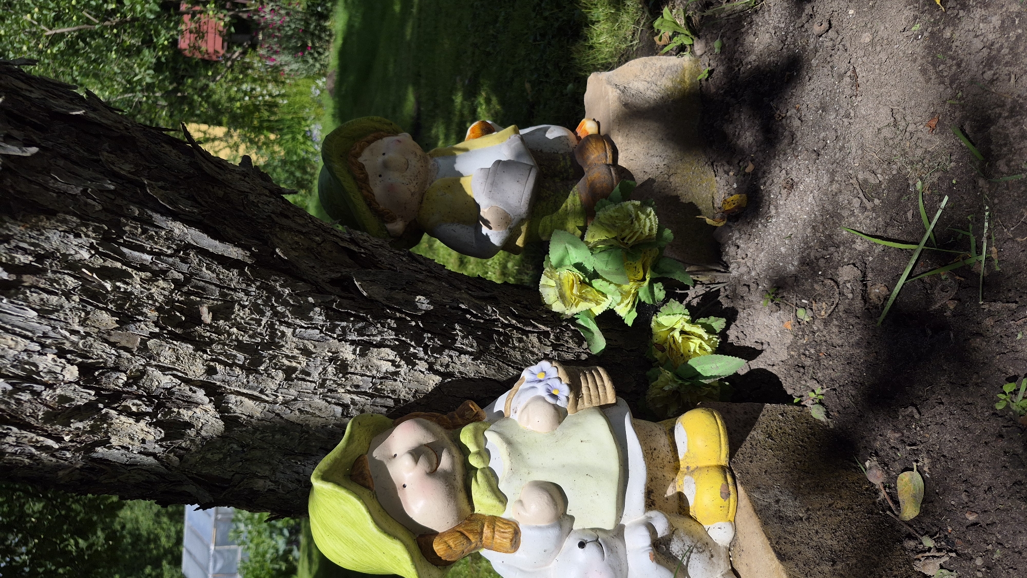 A frontal medium shot captures two garden gnome figurines positioned next to the trunk of a mature tree. The gnome on the left, dressed in a light green hat and apron over a white outfit, holds a basket with blue flowers. A white rabbit figurine is partially visible to its left. The gnome on the right wears a green hat and yellow vest over a white shirt, holding a grey bucket. Both gnomes have tan skin and dark hair. They are situated on dark soil, with a few artificial yellow flowers placed between them. The tree trunk is textured and dark. In the background, a well-maintained lawn is visible, along with some distant buildings and greenery. The scene is illuminated by sunlight, casting shadows on the ground.