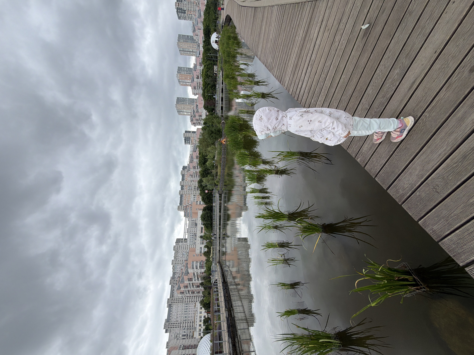 The image shows a cloudy day in a park. A young child in a hooded jacket and light-colored pants stands on a wooden walkway overlooking a calm body of water. The water reflects the cloudy sky and some reeds growing within it. In the background, across the water, are modern apartment buildings, characteristic of the architecture found in the city of Khimki, Russia. The walkway curves to the right, suggesting a park setting designed for walking and enjoying the scenery. The overall atmosphere is subdued due to the overcast weather.