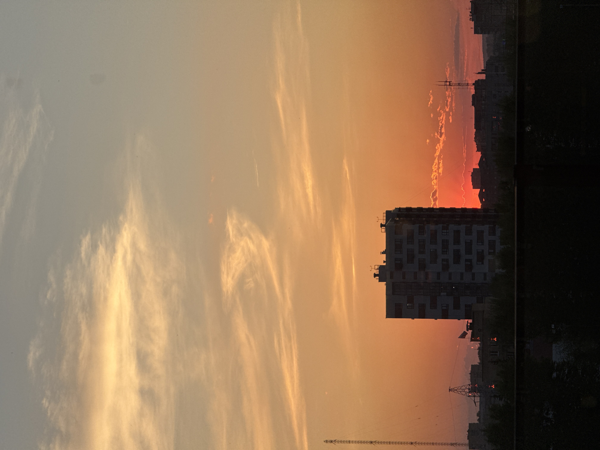 The image shows a sunset in Krasnoyarsk, Russia. The sky is filled with orange and yellow hues, and wispy clouds are visible. A tall apartment building is silhouetted against the colorful sky, along with other buildings in the distance. A construction crane is also visible, and the right side of the image is dark, possibly depicting trees or another building casting a shadow.