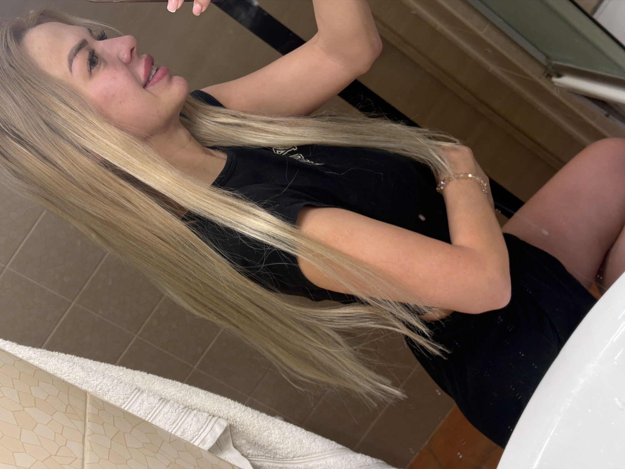 A young woman with long blonde hair, wearing a black t-shirt, is taking a selfie in a bathroom. She is smiling and looking at her reflection in the mirror. Her right hand is raised, holding a phone. A white towel is hanging on the wall behind her, and the walls are tiled with a beige, textured pattern. The scene appears to be in a residential bathroom. There are no identifiable landmarks or clues to indicate the specific location within Kyiv, Ukraine.