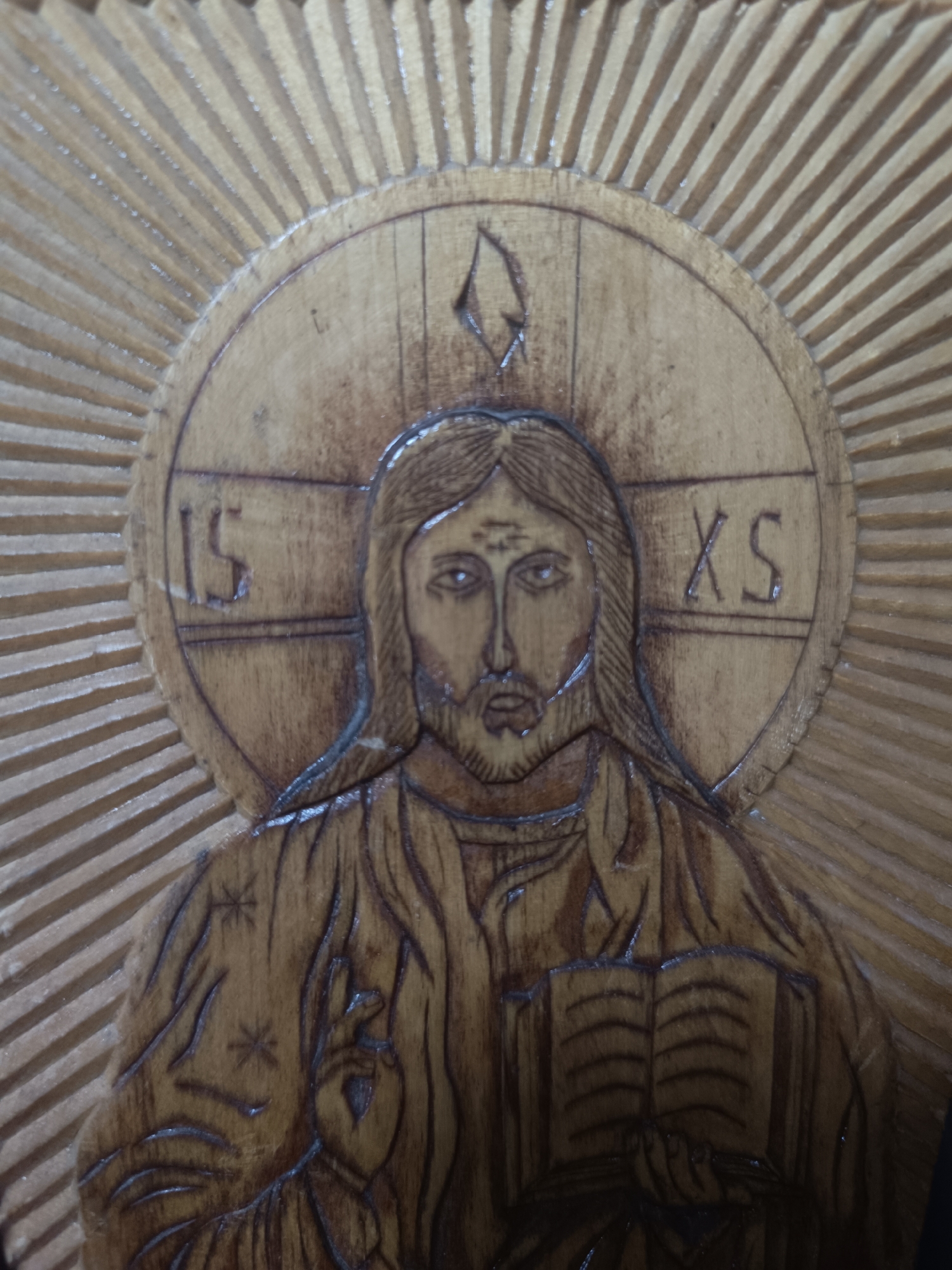 The image shows a carved wooden icon of Jesus Christ. He is depicted with long hair and a beard, holding a book in his left hand and making a gesture of blessing with his right. Around his head is a halo with the letters "IS" and "XS". Above his head, within the halo, there is a symbol resembling a flame. The halo is surrounded by radiating lines, suggesting light or glory. The carving style is relatively simple and rustic.
There is no information in the image to identify the city of Lermontov, Russia.