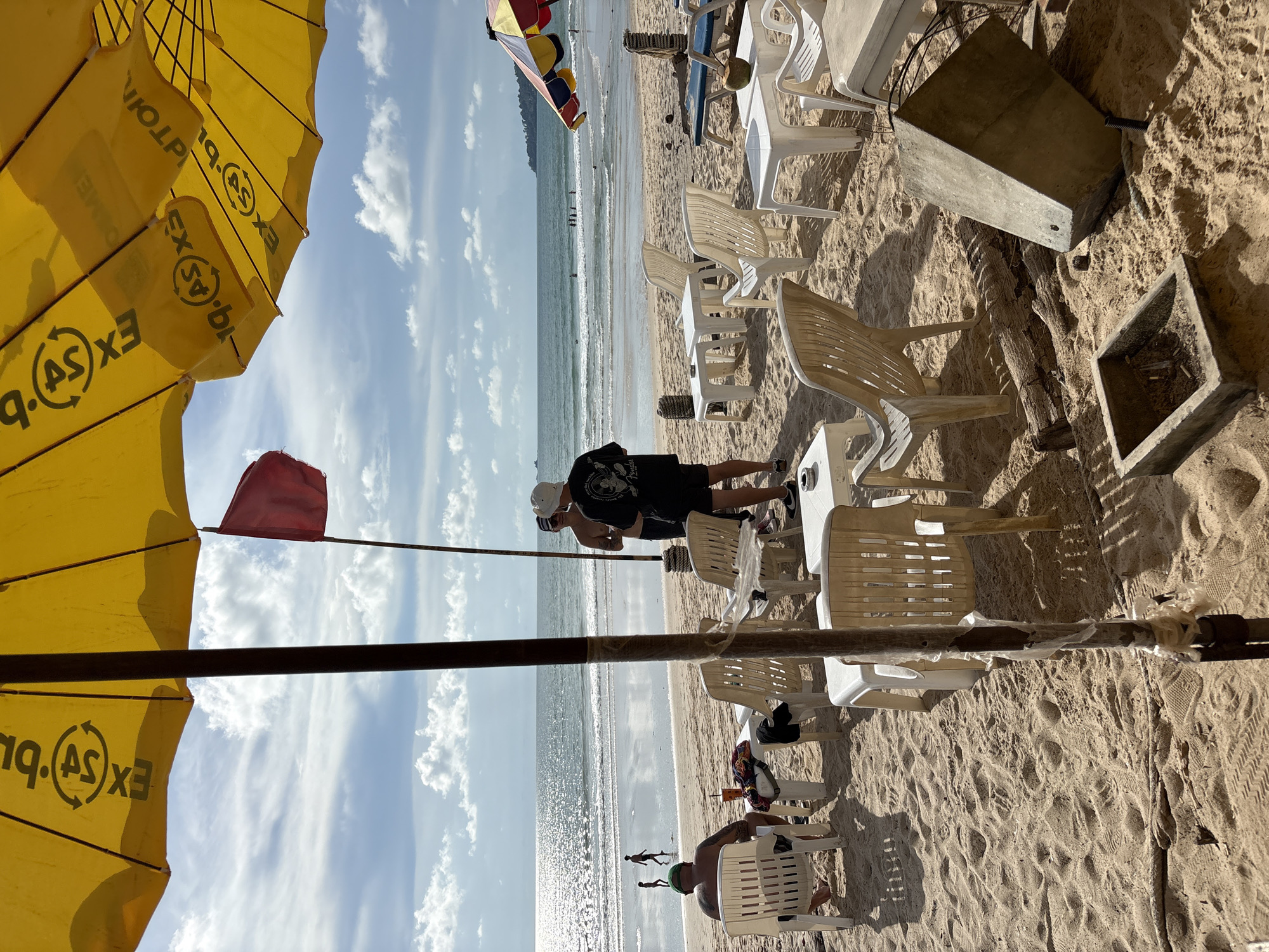 The image depicts a beach scene on a sunny day with a cloudy sky, likely in Choeng Thale, Thailand, given the presence of beach umbrellas and chairs commonly found in tourist areas. In the foreground, a yellow beach umbrella partially obscures the view, with visible branding "EXO" and "BUS". Scattered across the sandy beach are several white plastic chairs and small white tables, some arranged in seating areas. A red flag is planted in the sand behind a pole. Two men stand near the umbrella, facing away from the camera. One man wears a white hat and black t-shirt with a graphic design, and the other wears a dark t-shirt and a white baseball cap. Further back, a person sits in a white chair, facing the ocean, with a drink and a colorful patterned cloth on the table. In the middle ground, the ocean is visible with gentle waves and a few people wading or swimming in the distance. Two figures are walking on the wet sand near the water's edge. In the lower right corner, a large concrete object and a small concrete ash tray filled with cigarette butts are on the sand, alongside some weathered logs. The overall scene conveys a relaxed, outdoor atmosphere typical of a beach resort.
