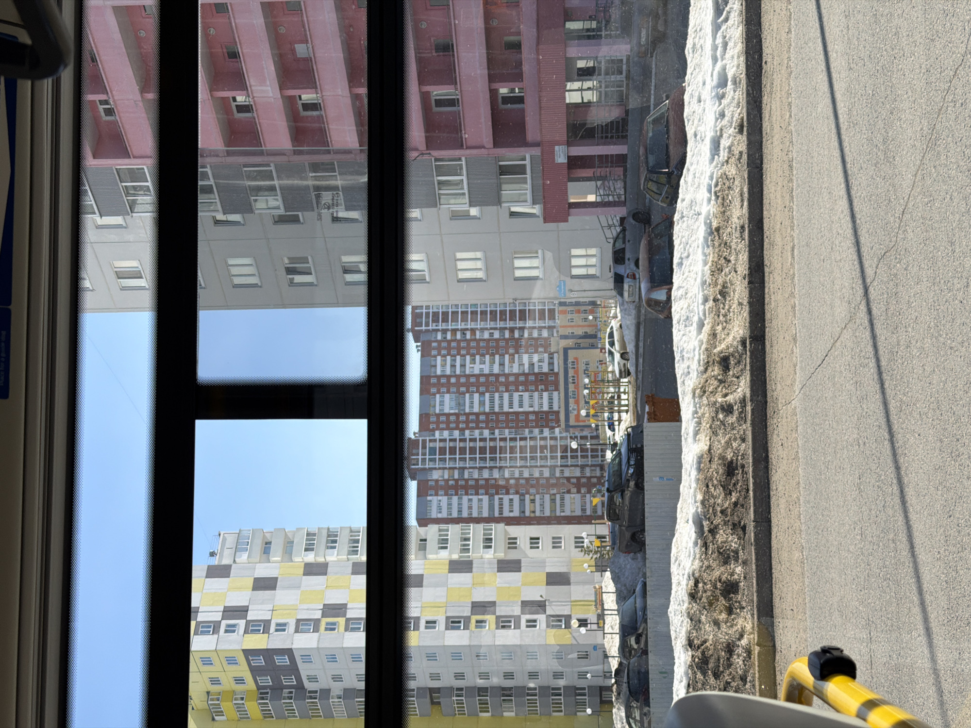 The image is a view from inside a vehicle, likely a bus or tram, through a window with multiple panes. Outside, the scene is urban with several multi-story buildings visible, reflecting in the window. The buildings vary in color and design, including pink, gray, and a yellow/gray checkered pattern. A tall red-and-white building is also visible. A row of parked cars lines the side of the road, next to a curb. Snow and dirty snowdrifts are piled up beside the road. The street appears to be asphalt. The location is Berezniki, Russia, indicated by the architectural style of the buildings, presence of snow, and urban layout. No people are visible in the shot.