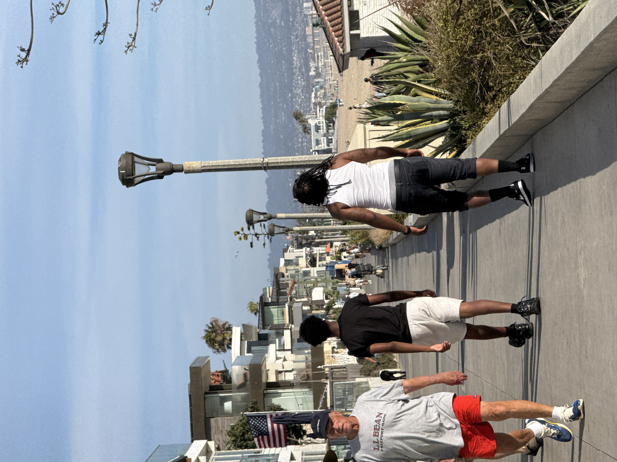 An eye-level, full-shot perspective captures a sunlit boardwalk in Manhattan Beach, California. Three men are walking towards the right foreground, two from the left and one from the right. The man on the far left is wearing a grey t-shirt with "LLBEAN FREEPORT MAINE" printed on it, red shorts, and blue and white sneakers. He has a blue baseball cap on. Next to him, walking slightly behind, is a young Black man in a black t-shirt and light grey shorts, black athletic shoes. To the right of him, and slightly ahead, is another man wearing a white tank top, dark knee-length shorts, and black athletic shoes. His hair is in dreadlocks.
The boardwalk is paved with concrete and extends into the background, lined by modern, multi-story residential buildings on the left and dense, spiky green plants and trees on the right. In the far distance, a hazy cityscape with buildings and hills is visible under a pale blue sky with scattered clouds. A lamppost stands prominently on the right side of the boardwalk. The overall scene depicts a casual, outdoor environment with people enjoying a walk along the coast.