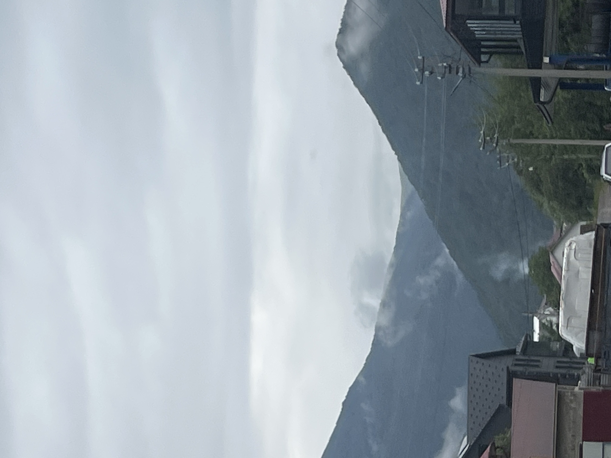 The image shows a cloudy sky over a mountainous landscape in the village of Krasna, Ukraine. In the foreground, there are several buildings and a street. On the left, a building with a dark grey roof and a brown-red lower section is visible. Adjacent to it is another dark grey building with multiple windows. In the center, a white object, possibly a vehicle or structure, is parked on a dark surface, and a small yellow figure is seen near it. Further to the right, a building with a dark pitched roof and glass windows is present. Utility poles with wires run through the scene. In the background, the lush green slopes of mountains are covered in parts by mist and clouds. Smoke plumes are visible rising from the mountainside, suggesting human activity such as heating or cooking. The overall atmosphere is overcast and slightly hazy.