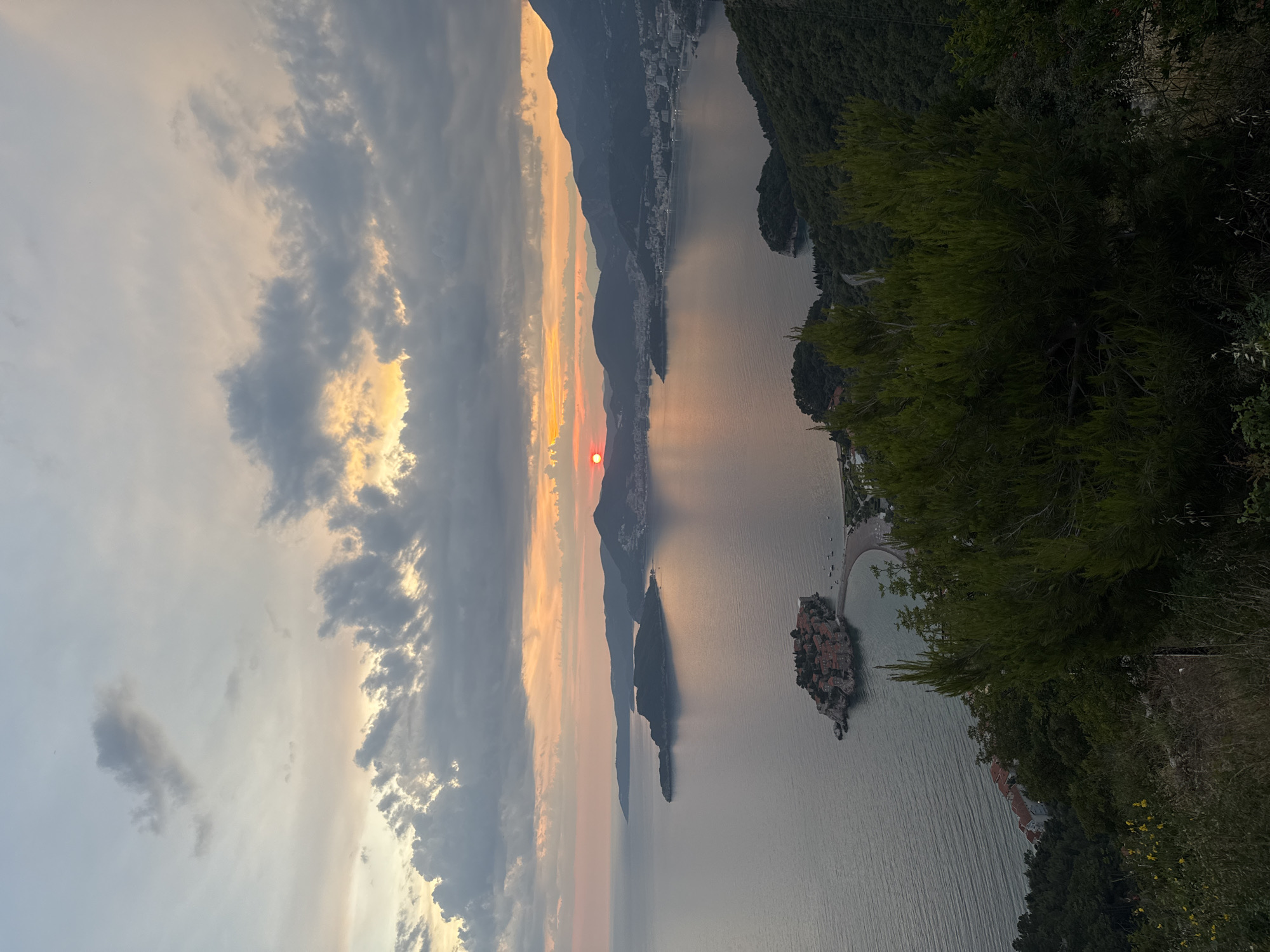 This image captures a panoramic view of the island town of Sveti Stefan, Montenegro, during a sunset. The sky is filled with a gradient of colors, transitioning from soft oranges and yellows near the horizon to dusky blues and grays higher up. Dramatic clouds, tinged with the setting sun's light, drift across the sky.
The island of Sveti Stefan, a cluster of red-roofed buildings connected to the mainland by a narrow causeway, is prominently featured in the foreground. The calm, reflective waters of the Adriatic Sea surround the island. Beyond Sveti Stefan, the coastline curves, revealing a bay dotted with buildings and backed by rolling, tree-covered hills. The sun, a distinct red orb, is low on the horizon, casting a warm glow over the scene. In the immediate foreground, lush green trees and foliage obscure the lower portion of the view, creating a sense of elevation and framing the wider landscape. No people or discernible actions are present in the image.