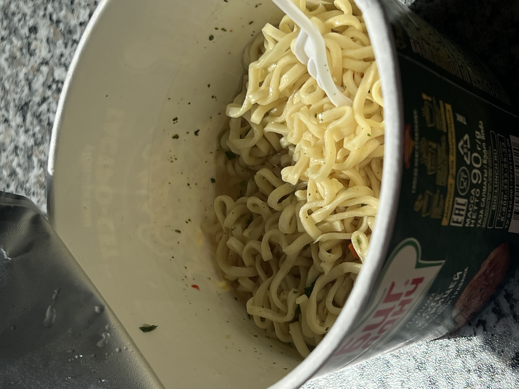 The image shows a close-up of a cup of instant noodles. The cup is white and has a green label with some text and images. The noodles are cooked and submerged in broth. A white plastic fork is visible in the noodles. The background is a speckled surface, possibly a countertop. There is no indication of Zelenodol'sk, Russia, or any people in the image.