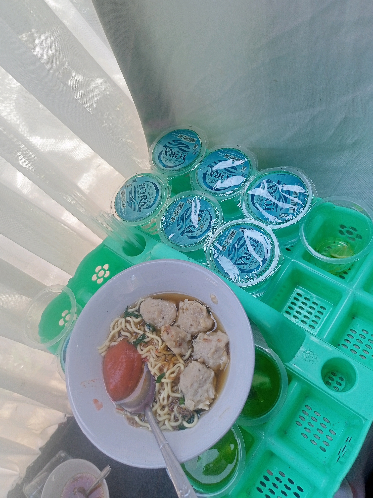 The image depicts a table setting with food and drinks, likely a meal being prepared or served, possibly at a street food stall in Limbangan, Indonesia. In the foreground, a white bowl contains a portion of noodles, meatballs, and a large piece of tomato in broth, with a silver spoon resting in the bowl. To the right of the bowl is a green plastic tray with multiple compartments. Several small, clear plastic cups containing a green liquid are visible within some of these compartments. Above the tray, a cluster of small, clear plastic cups filled with mineral water, branded "YORA," are arranged. The background features a white, sheer curtain, allowing diffused light to enter, creating a soft and somewhat hazy atmosphere. The overall scene suggests an outdoor dining setup, common in such Indonesian locales.