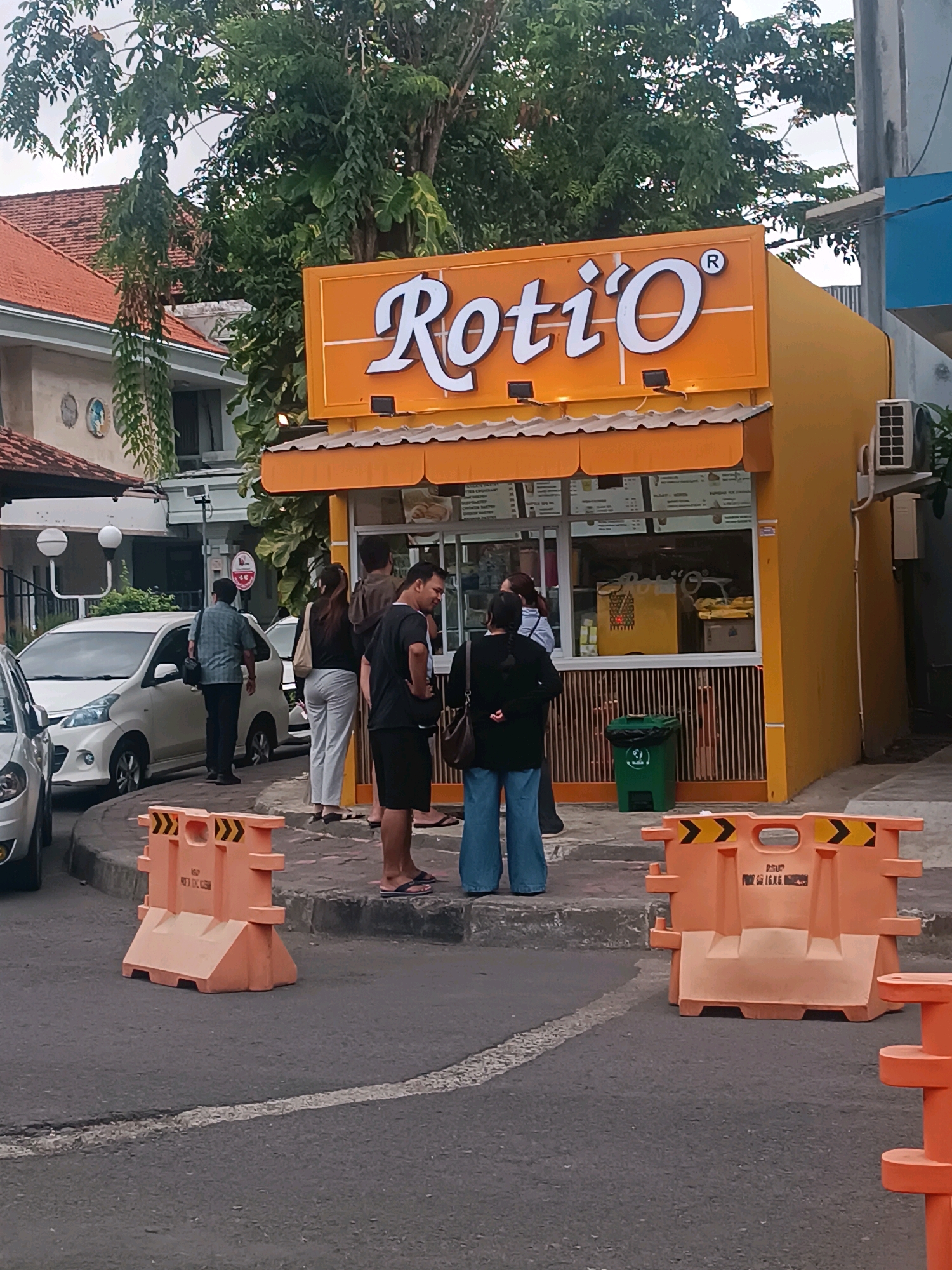 The image depicts a street scene in Denpasar, Indonesia, featuring a small, orange food stall named "Roti O". The stall has a prominent sign with white lettering. Several people are gathered in front of and around the stall. Two individuals stand closest to the camera, one wearing blue jeans and a dark top, and another in a dark shirt and shorts. Further back, a group of four people are visible, one in a checkered shirt, another in a dark top and white pants, a person in a dark top and jeans, and a person in a dark shirt. A white car is parked to the left of the stall, and another white car is partially visible behind it. Orange traffic barriers are placed on the street in the foreground and mid-ground. A green trash can is situated near the stall. The environment includes a building with a red-tiled roof in the background and lush green trees overhead. The overall scene appears to be a typical urban setting with commercial activity.