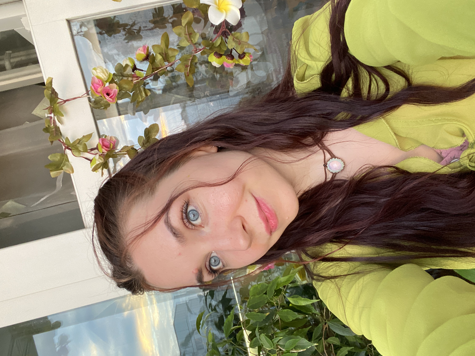 The media file shows a close-up, selfie-style shot of a young woman with long, dark, wavy hair and striking blue eyes. She is wearing a light green, flowy garment and a delicate necklace with a pale, oval pendant. Her expression is gentle, with a slight smile and her eyes looking towards the camera.
Behind her, a decorative vine with pink and white artificial flowers is draped across a white frame, possibly a window or door. Reflections in the glass suggest an outdoor setting, with what appears to be a sunset or sunrise casting warm light, indicating either early morning or late evening. Green foliage is visible in the lower portion of the frame, further reinforcing an outdoor or garden-like environment. The overall mood is serene and pleasant. There is no visible text in the image. The location context provided is Chaikovsky, Russia.