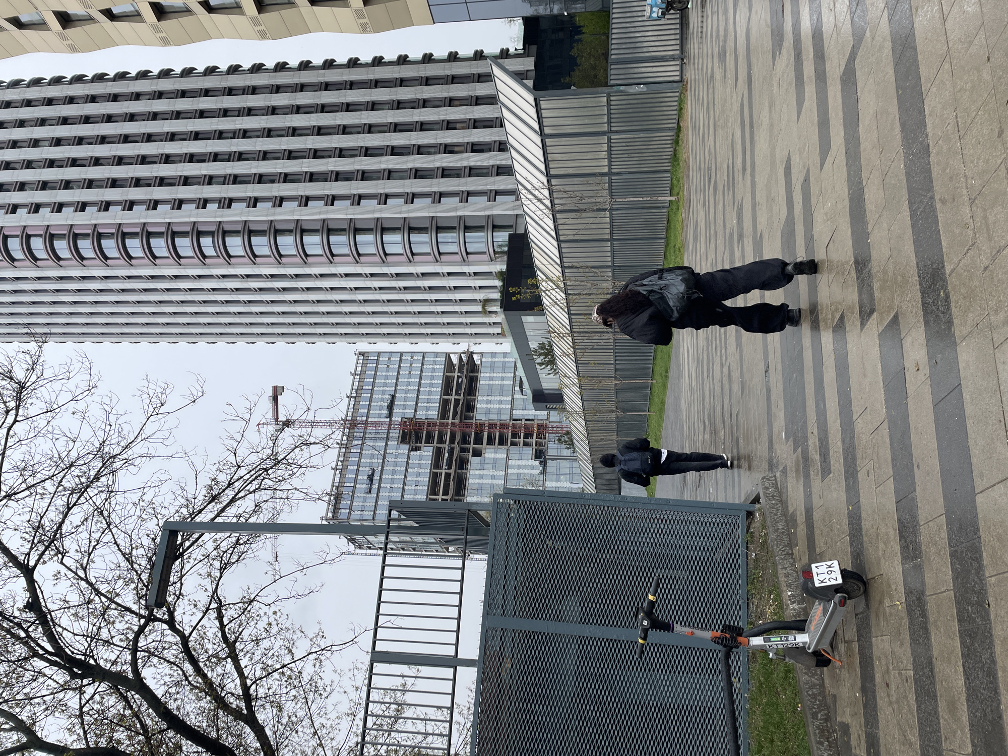 The image shows an outdoor scene in Khimki, Russia. Two individuals are walking along a paved walkway. A tall building with a modern facade of repeating windows dominates the background on the left. A building under construction with a crane is visible behind a metal fence. A scooter with a license plate reading "KT1 29K" is parked near the fence. The weather appears overcast. Bare trees are visible in the foreground.