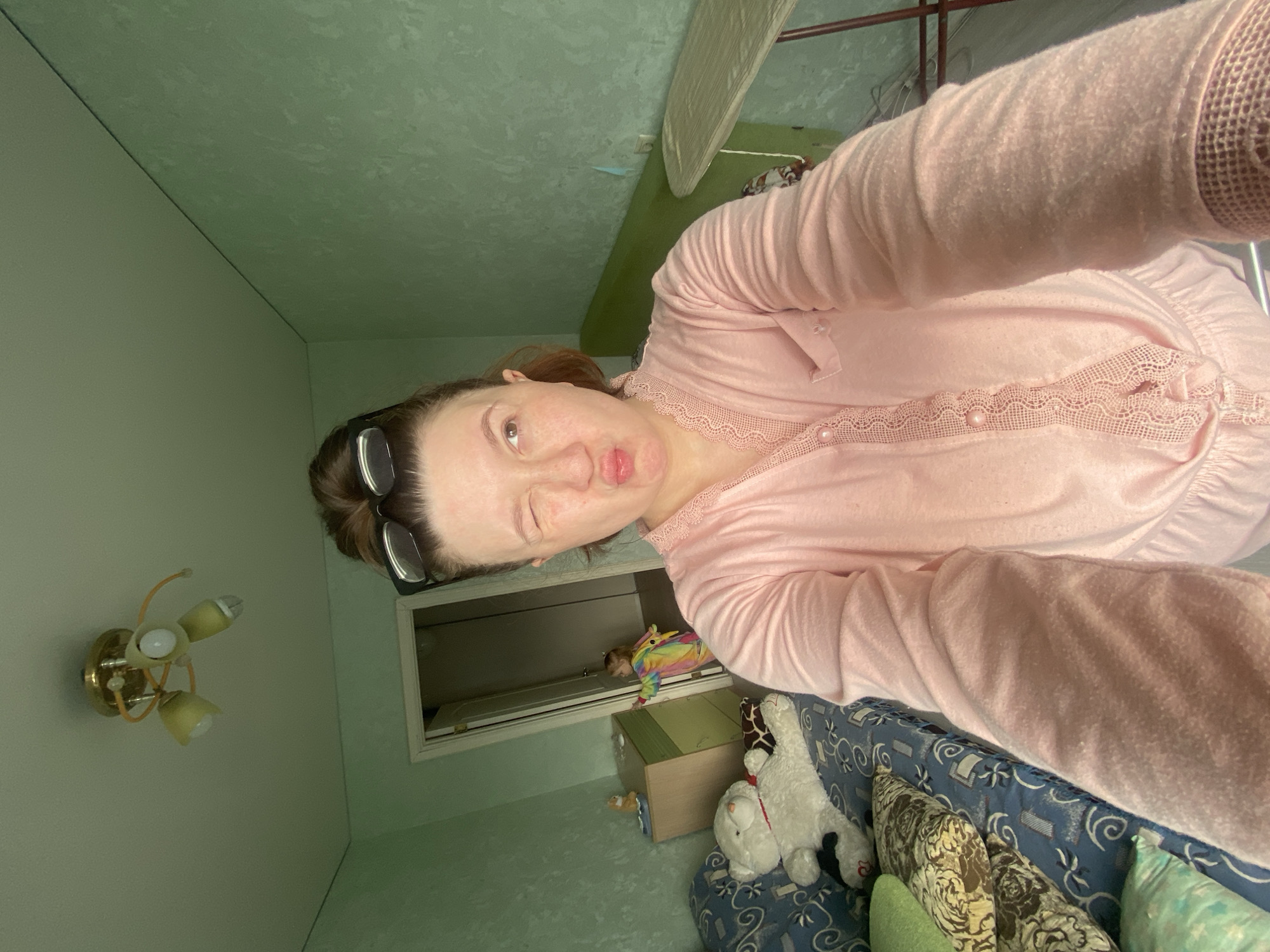 An adult female, estimated to be in her 30s-40s, is captured in a selfie taken indoors, likely in Chaikovsky, Russia, though the precise location within the city is undetermined. She has light skin, light brown hair pulled back, and black-framed eyeglasses resting on her forehead. She is wearing a light pink, long-sleeved top featuring lace detailing and pearl-like buttons. Her right eye is closed in a wink, and her lips are puckered. The camera is held by her outstretched right arm, partially visible in the lower right.
The room has light green, textured wallpaper. A brass-colored ceiling fixture with three light bulbs, two with shades and one exposed, is mounted on the light-colored ceiling. To the left, a blue sofa with a white and blue patterned upholstery is present, holding a large white teddy bear with a red ribbon and multiple pillows, including a green one and two with brown floral patterns. Behind the teddy bear, a smaller, possibly giraffe-themed, stuffed animal is visible. Adjacent to the sofa is a light wood dresser with two drawers, one of which is light green. A small orange-brown figurine and a blue object rest on top of the dresser. An open white doorway in the background reveals a young child, wearing a brightly colored, possibly rainbow-themed, hooded garment, looking into the room. On the far right, a light green ironing board stands upright on metal legs.