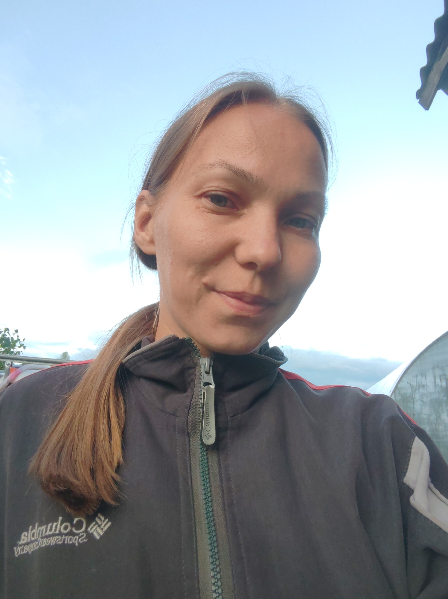 A medium shot captured from chest-up shows a woman from the chest up, facing the camera and smiling faintly. She has light skin, light brown hair tied back in a ponytail, and blue eyes. She is wearing a dark gray jacket with red and white accents on the shoulders and a zipper down the front. The jacket has a Columbia logo on the chest. The background is a light blue sky with scattered clouds. To the right of the woman, a portion of a white, curved structure, possibly a greenhouse, is visible. The overall scene appears to be outdoors, in an environment that might be rural or on the outskirts of a city. The image has a slightly overcast lighting condition.