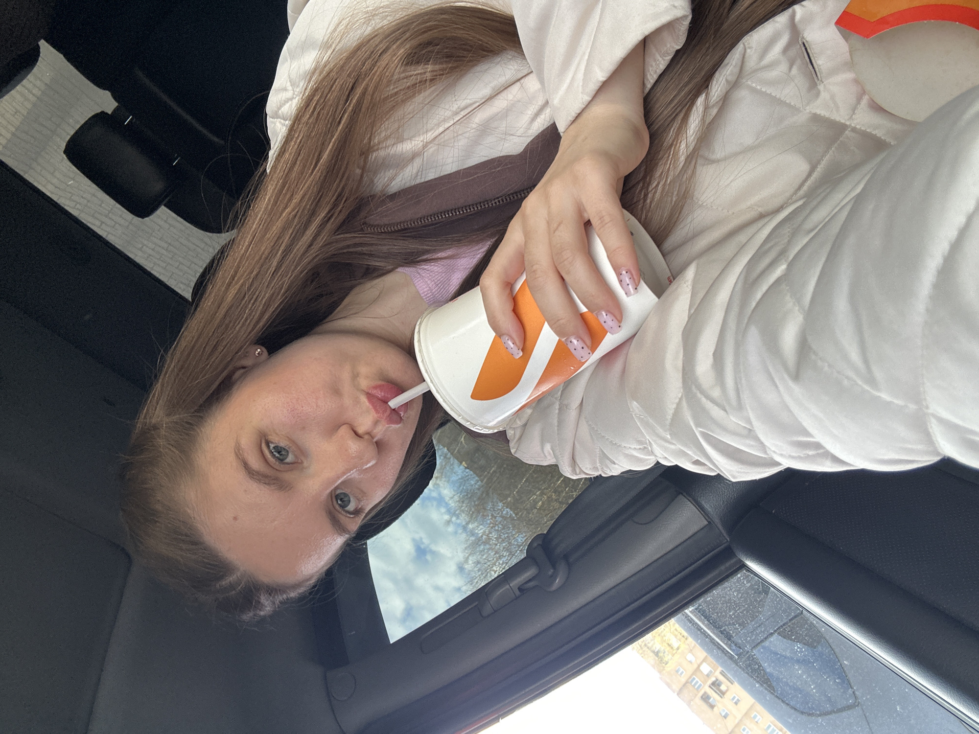 A woman is photographed inside a car, holding a cup with an orange logo and sipping from a straw. She is wearing a light-colored, quilted jacket over a brown and pink top. Her long, brown hair falls to one side. The car's interior is visible, with the roof open, revealing a view of a building, possibly located in the city of Klin, Russia.