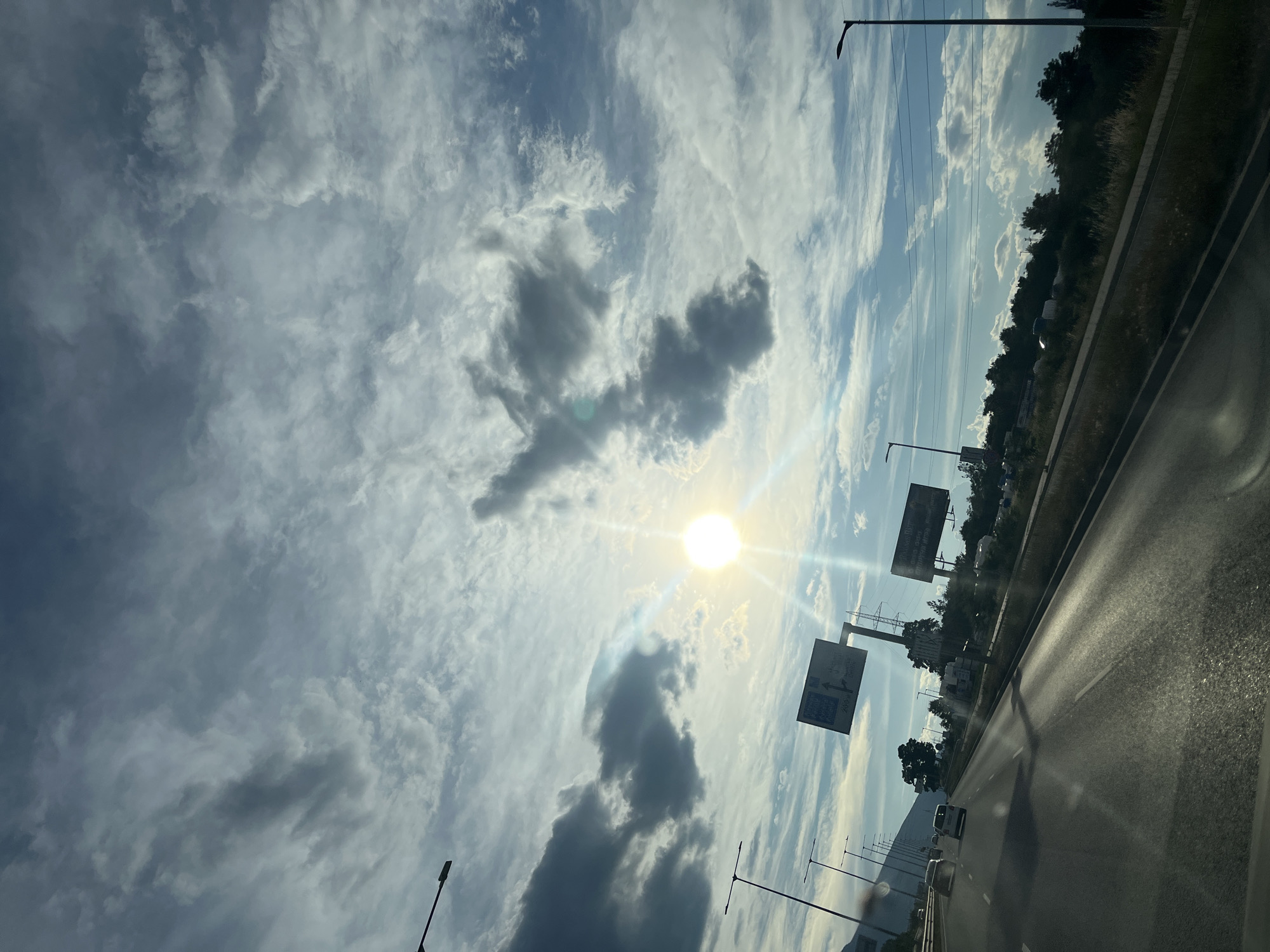 The image shows a view from a vehicle traveling on a highway. The sun is visible in the sky, shining brightly through scattered clouds, suggesting it is daytime. The sky is a mix of blue and white, with some darker clouds present. On the right side of the image, there is a highway with multiple lanes and other vehicles. Along the side of the highway, there are trees and roadside infrastructure, including streetlights and large billboards. The billboards have some text on them, but it is not legible at this resolution. The overall scene appears to be an outdoor, daytime view of a road journey. The lighting and cloud patterns suggest a bright but possibly dynamic weather day. The perspective indicates the photo was taken from the driver's or a passenger's seat.