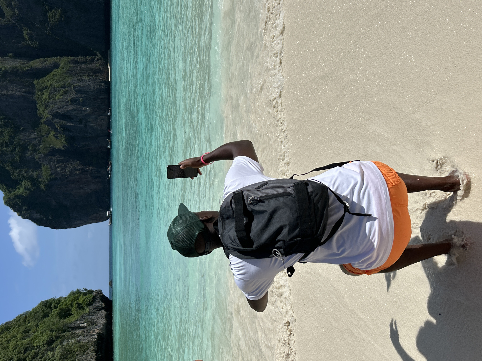 A person wearing a white t-shirt, orange shorts, and a green baseball cap kneels on a sandy beach, holding a smartphone to take a picture. A black backpack is worn over the t-shirt. The water in front of the person is a vibrant turquoise, and large limestone cliffs covered in greenery rise from the sea in the background. Several boats are visible in the distance. This scene is characteristic of the beaches in Ao Nang, Thailand, known for their dramatic karst topography and clear waters. The sun is shining, creating bright light and shadows on the sand.