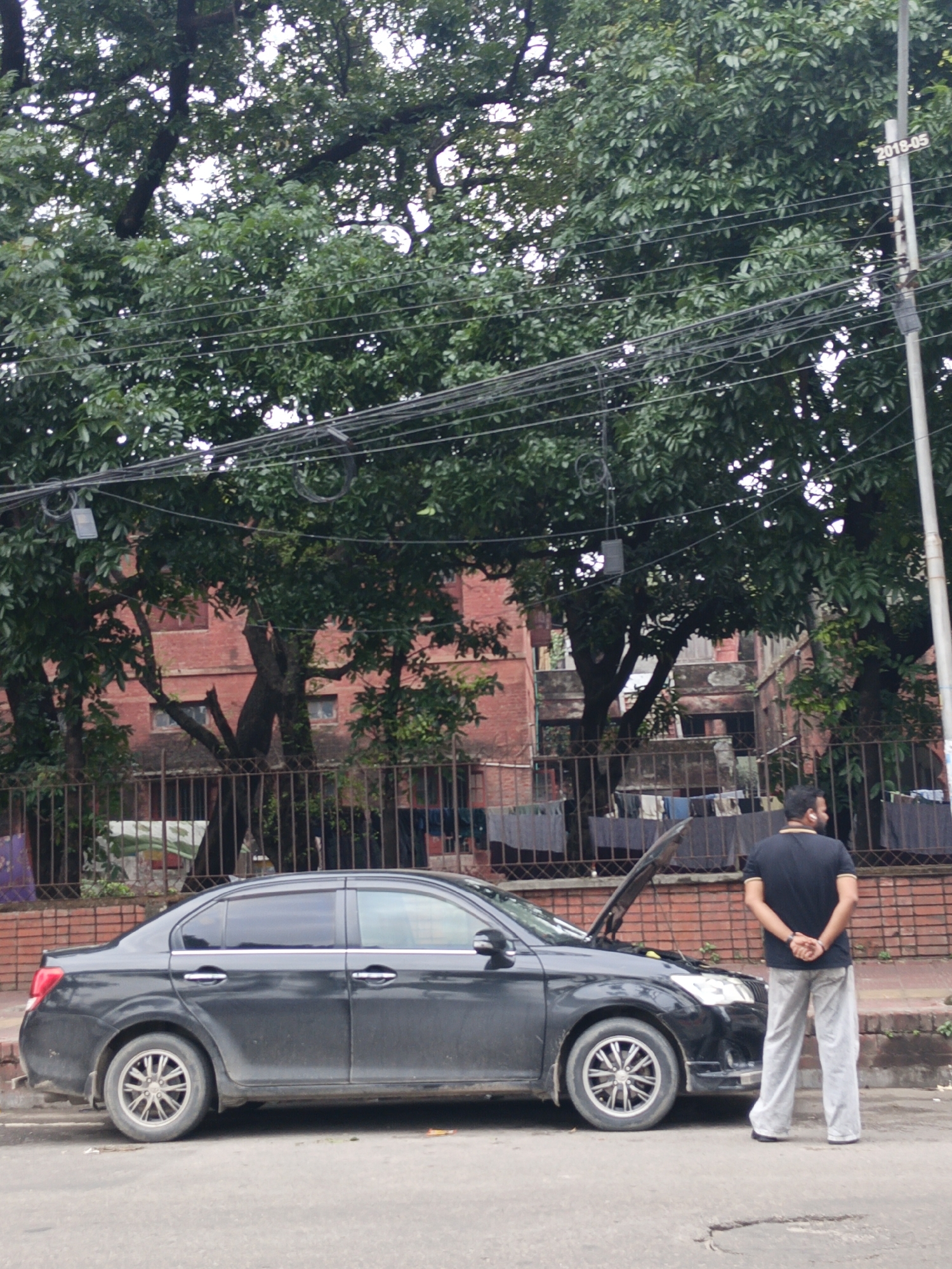 The image depicts an outdoor urban or semi-urban scene during the daytime, likely under overcast weather conditions as suggested by the diffused lighting.
The primary subjects are a man and a black sedan car. The car is parked on the side of a road or sidewalk, with its hood open, indicating a potential breakdown or ongoing maintenance. A man stands at the front-right side of the car, with his back to the camera. He is wearing a dark t-shirt and light grey or white pants, and appears to have white earbuds in his ears. His hands are clasped behind his back, suggesting a posture of waiting or observation.
In the background, a tall electricity pole is visible on the right, bearing the text "2018-05". A dense network of overhead utility lines stretches across the upper-middle section of the frame. Behind the car and the man, there is a metal fence, with clothes hanging on a line or drying rack visible behind it. Further back, large trees with lush green foliage dominate the upper part of the image, partially obscuring several red brick buildings with windows. The scene conveys a moment of everyday life, possibly dealing with a car issue.