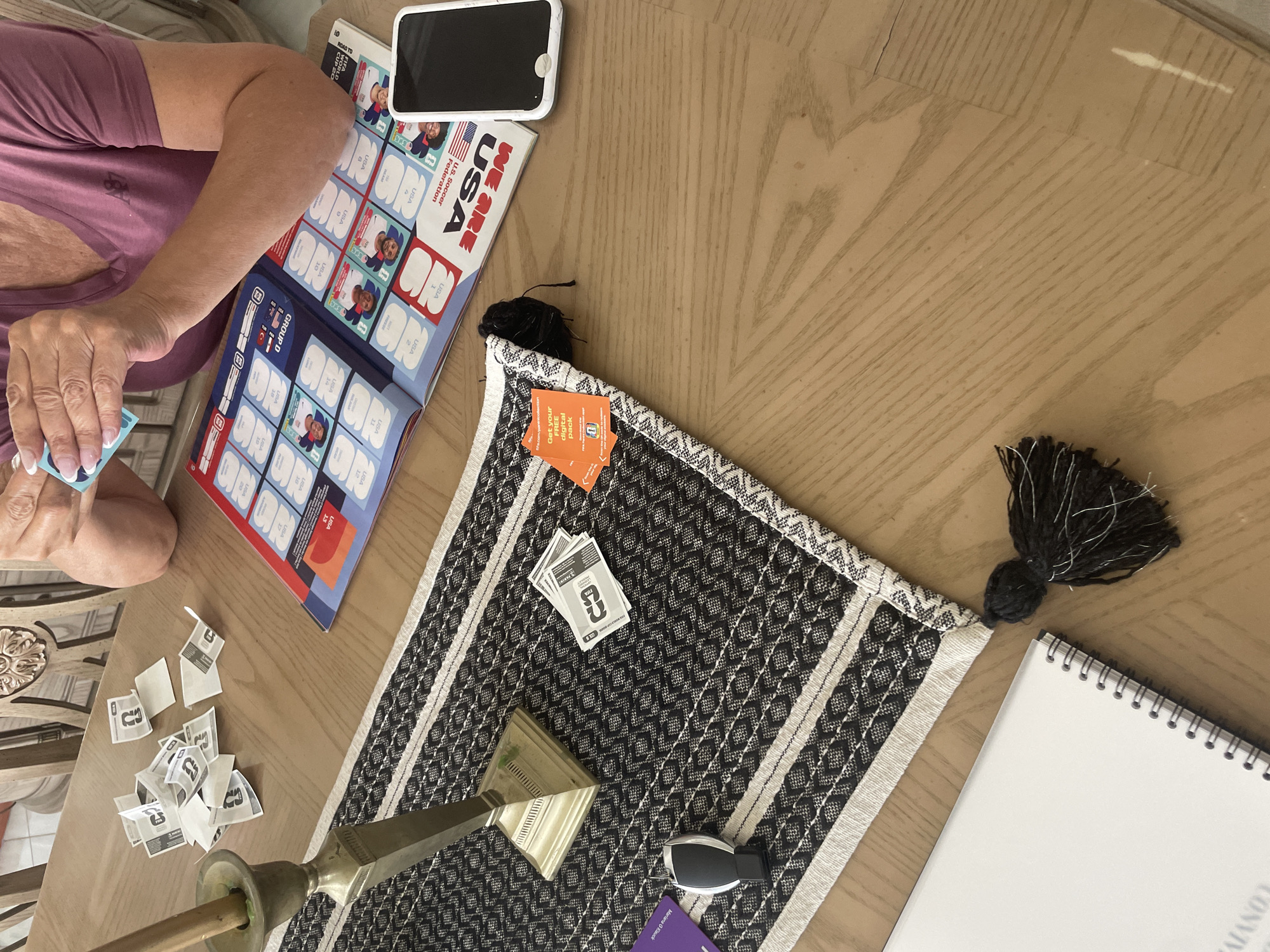 Here is a detailed description of the image:
The image shows a person sitting at a wooden table and participating in an activity involving stickers or cards from a "We Are USA" U.S. Soccer Federation book. The person is shown holding a sticker and appears to be placing it into the book. A smartphone is adjacent to the book. A stack of empty sticker backing sheets is scattered on the table near the book. A patterned black and white table runner lies across the table. There are additional cards on the table runner along with a decorative pyramid and car key. A notebook is located on the table runner to the right of the table runner.
Given the items in the image, it appears that this is a personal and informal setting. There is no indication of San Bernardino Tlaxcalancingo, Mexico.