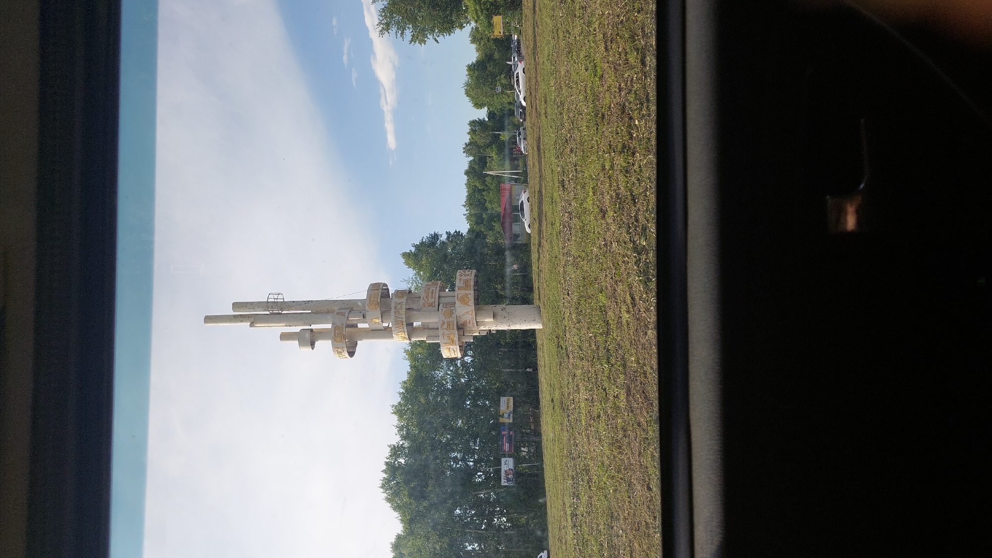 The image shows a monument in the city of Amursk, Russia, on a sunny day. The monument, made of concrete and metal, is tall and abstract, with spiraling elements and what appear to be decorative discs. It stands in a grassy field. In the background, there is a line of trees, several billboards, a building with a red roof, and a road with several cars parked and driving. The sky is blue with some white clouds. The photograph appears to be taken from inside a vehicle, as suggested by the dark interior framing the view.