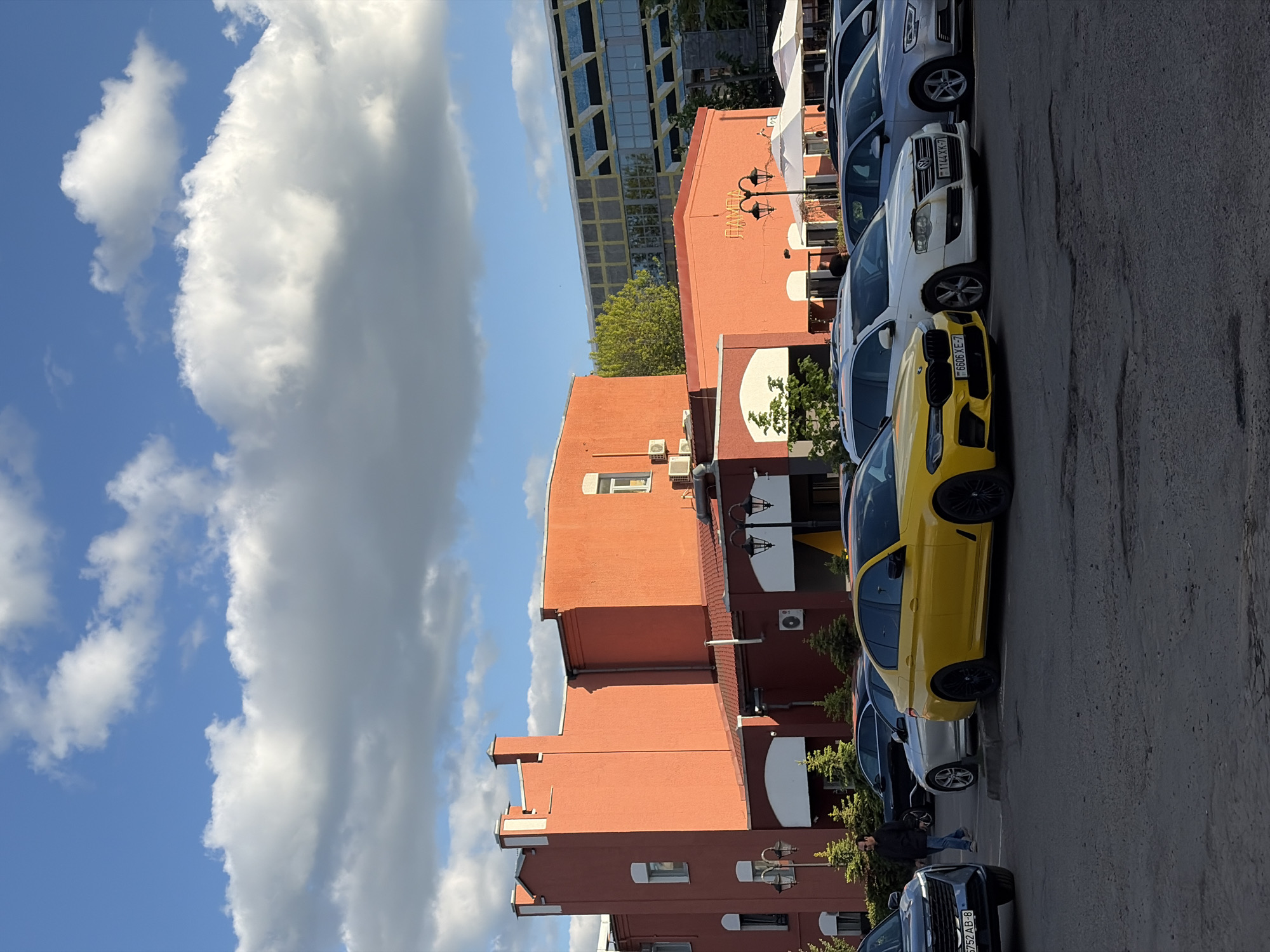 A street scene in Minsk, Belarus, features a row of parked cars including a bright yellow car. The building behind the cars is a reddish-brown brick structure with architectural details such as windows and air conditioning units. The sky is blue with puffy white clouds. A modern glass building is visible in the background. A person is walking near the parked cars.