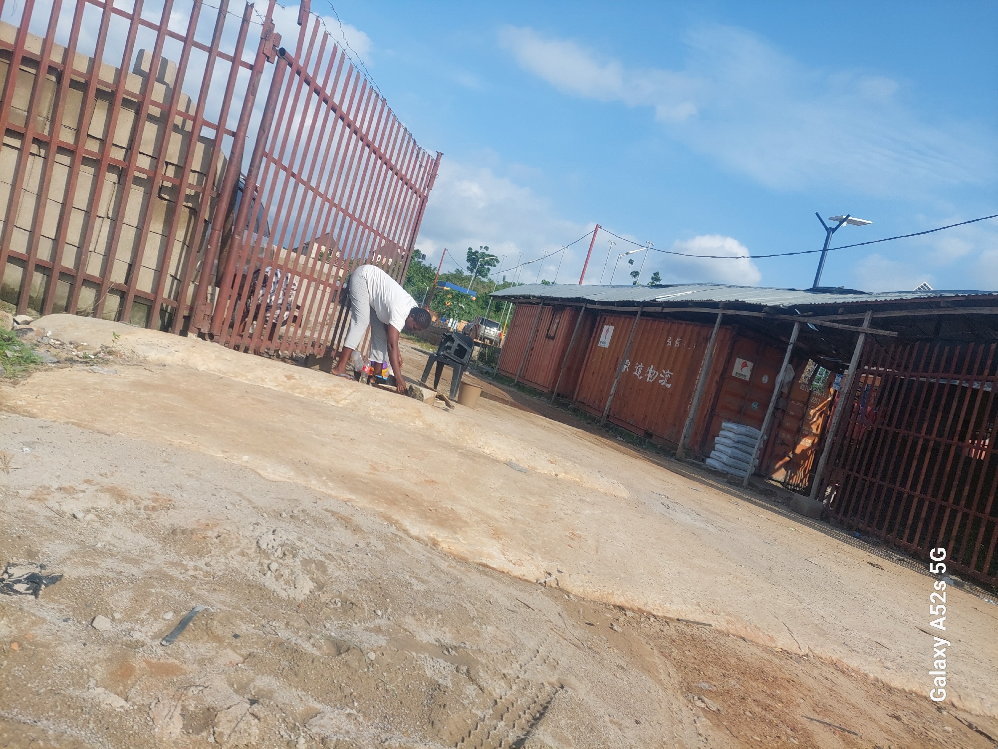 In Awkuzu, Nigeria, a person in a white shirt and light-colored pants is bending over near some shipping containers. The scene features a rough, sandy embankment on the left, a red metal fence or roof structure overhead, and the sky with scattered clouds. To the right, several large orange shipping containers are visible, some with markings. The ground appears uneven and possibly dusty.