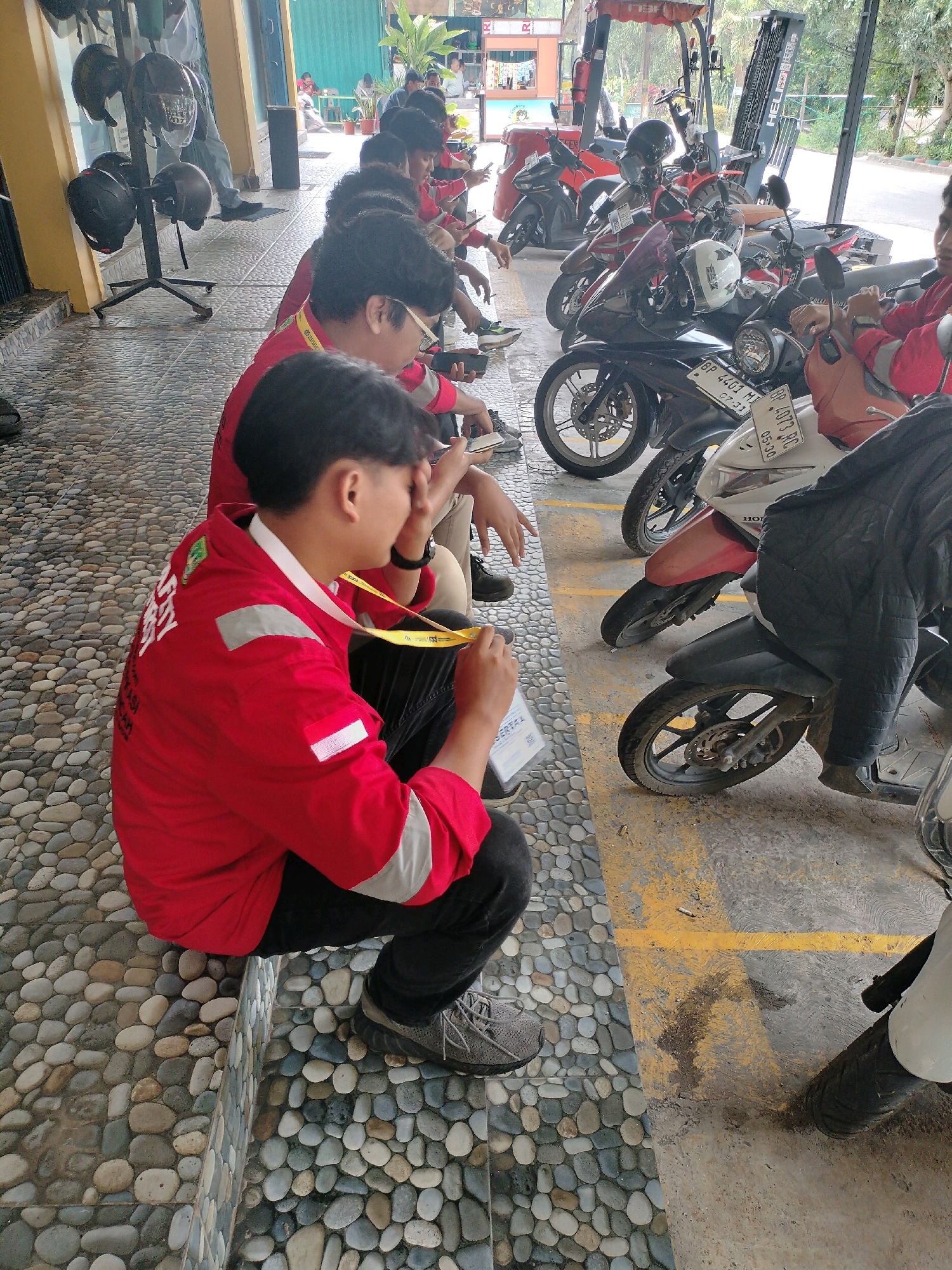The image shows a group of young men, likely students or workers, in a covered outdoor area, possibly a parking lot or waiting area, with a row of motorcycles parked behind them. The ground is paved with smooth, multicolored river stones. Several individuals are wearing red jackets, suggesting a uniform or team affiliation. One individual in the foreground is wearing a red jacket with white reflective stripes on the sleeves and a yellow lanyard around his neck. He is seated and holding a white piece of paper or a tablet. Other individuals are also seated or standing behind him, some looking at their phones. In the background, there are several parked motorcycles, a forklift, and what appears to be a building with windows. The overall atmosphere seems casual, possibly during a break or waiting period. The presence of motorcycles and the outdoor setting suggests an urban or semi-urban environment. While the specific location in Batam, Indonesia, cannot be definitively identified from the image alone, the style of the motorcycles and the general ambiance are consistent with common scenes in Indonesia.