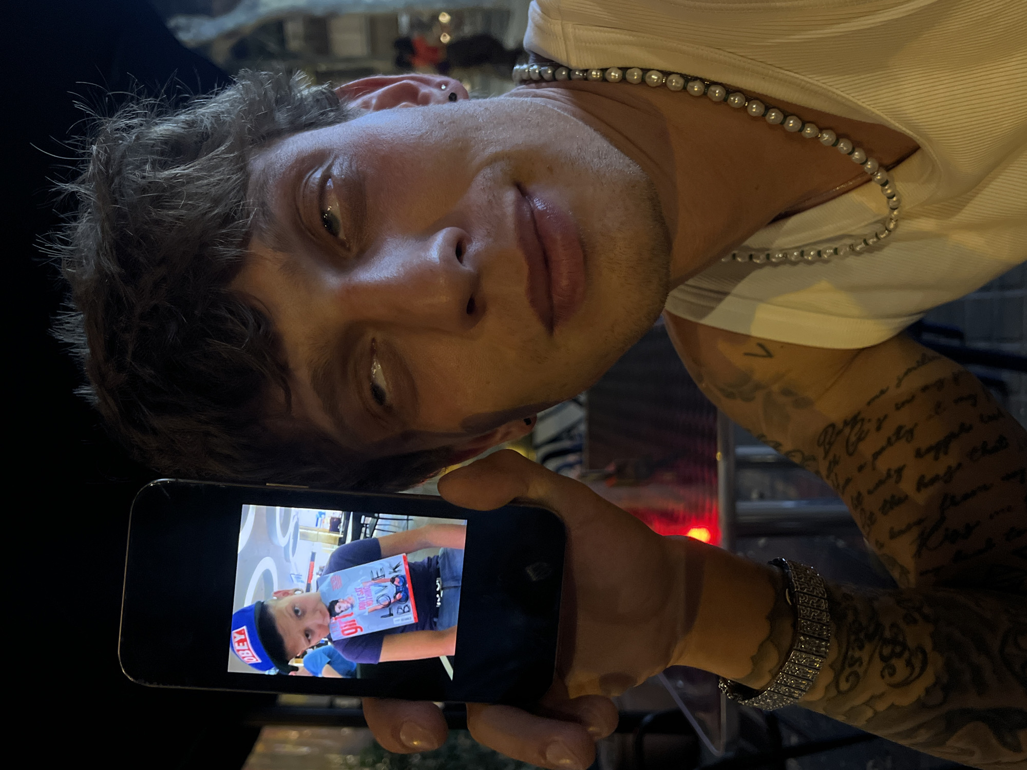 A close-up, outdoor, nighttime shot captures a man in the foreground holding a smartphone displaying a picture. The man has dark, messy hair and is looking up and to his right, with his mouth slightly open. He is wearing a white tank top and a pearl necklace. On his left wrist, he wears a diamond-studded bracelet. His arms are covered in tattoos. The smartphone screen shows a young person wearing a blue "OBEY" baseball cap, holding a magazine with the title "LOVE BOOK" and an image of a person on the cover. The background is blurred but suggests a restaurant or bar setting, with red lights visible. This scene appears to be in Barcelona, Spain, likely on a warm evening.