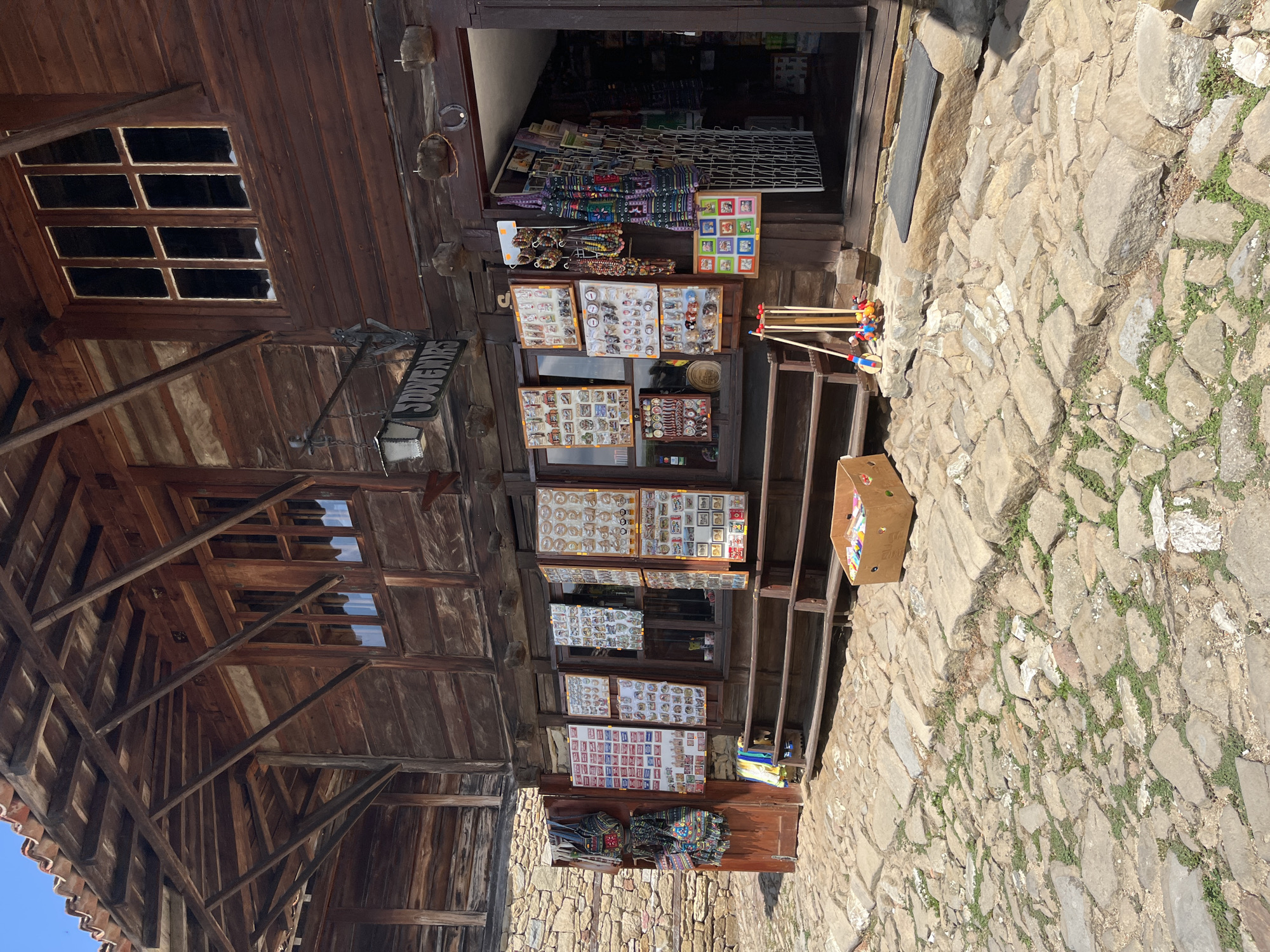 The image depicts the exterior of a souvenir shop in the historic town of Zheravna, Bulgaria. The shop is housed in a traditional wooden building with a tiled roof and exposed wooden beams. In front of the shop, various souvenirs are displayed on wooden boards and racks, including bags, jewelry, magnets, and postcards. A cobblestone street leads up to the shop entrance, which is open, revealing more merchandise inside. A cardboard box filled with colorful items is placed on the street in front of the shop. A simple wooden bench is situated in front of the display windows. The overall scene is representative of a charming, historical tourist attraction.