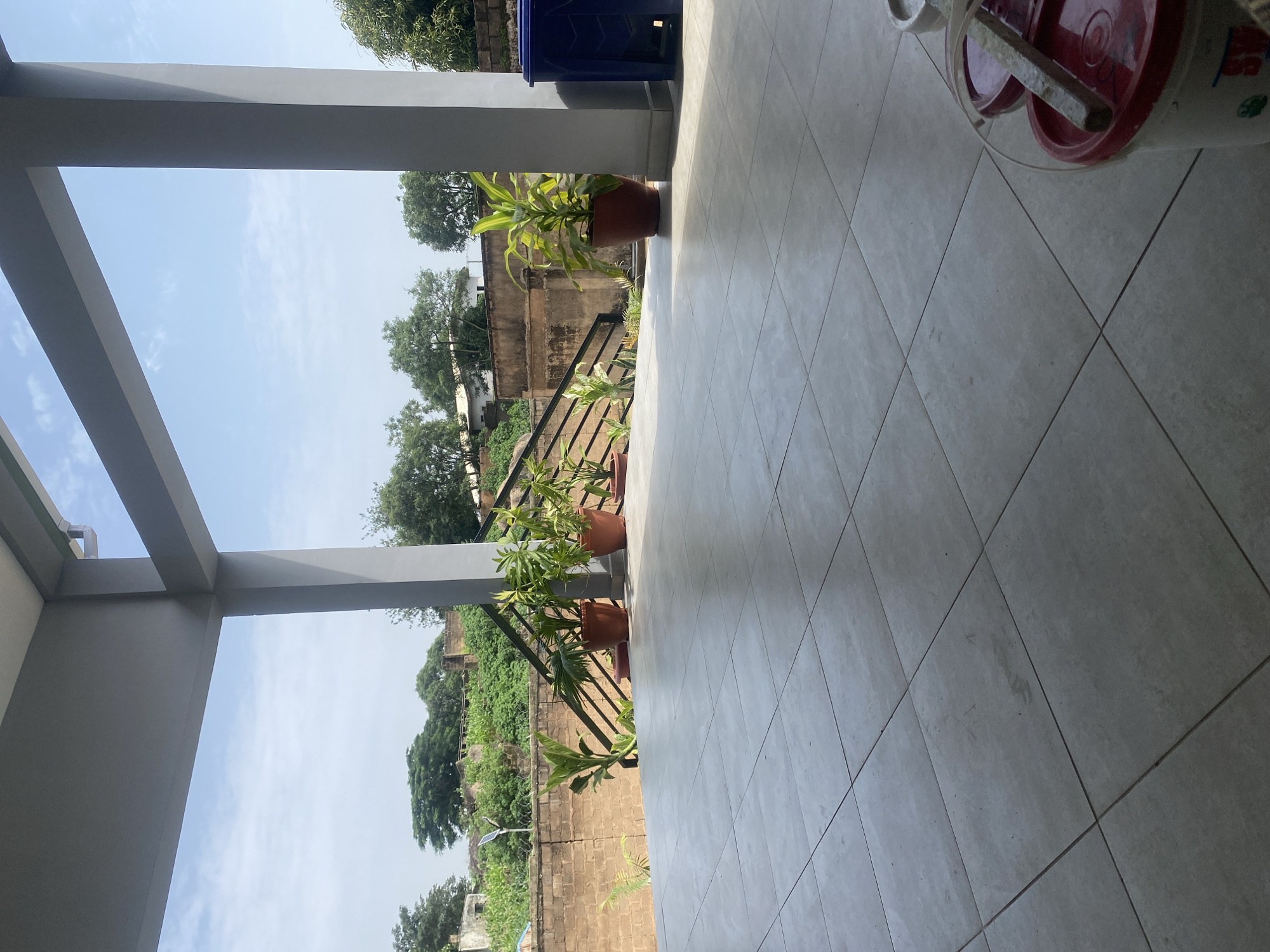 The image depicts a tiled patio area with a covered section supported by grey pillars and a ceiling. In the background, a brick wall separates the patio from an area with stairs leading up to a verdant hillside dotted with trees and some buildings. Several potted plants are arranged along the edge of the patio, including a large potted plant with broad green leaves in the foreground. To the right of the potted plants, a large blue bin is visible. The foreground is dominated by a light-colored tiled floor, with some buckets and a stick resting on the right side of the frame. The sky above is clear with scattered clouds. The scene appears to be an outdoor living space in a residential area. The mention of Jos, Nigeria suggests a specific geographical context, implying a likely warm climate and the presence of tropical or semi-tropical vegetation.