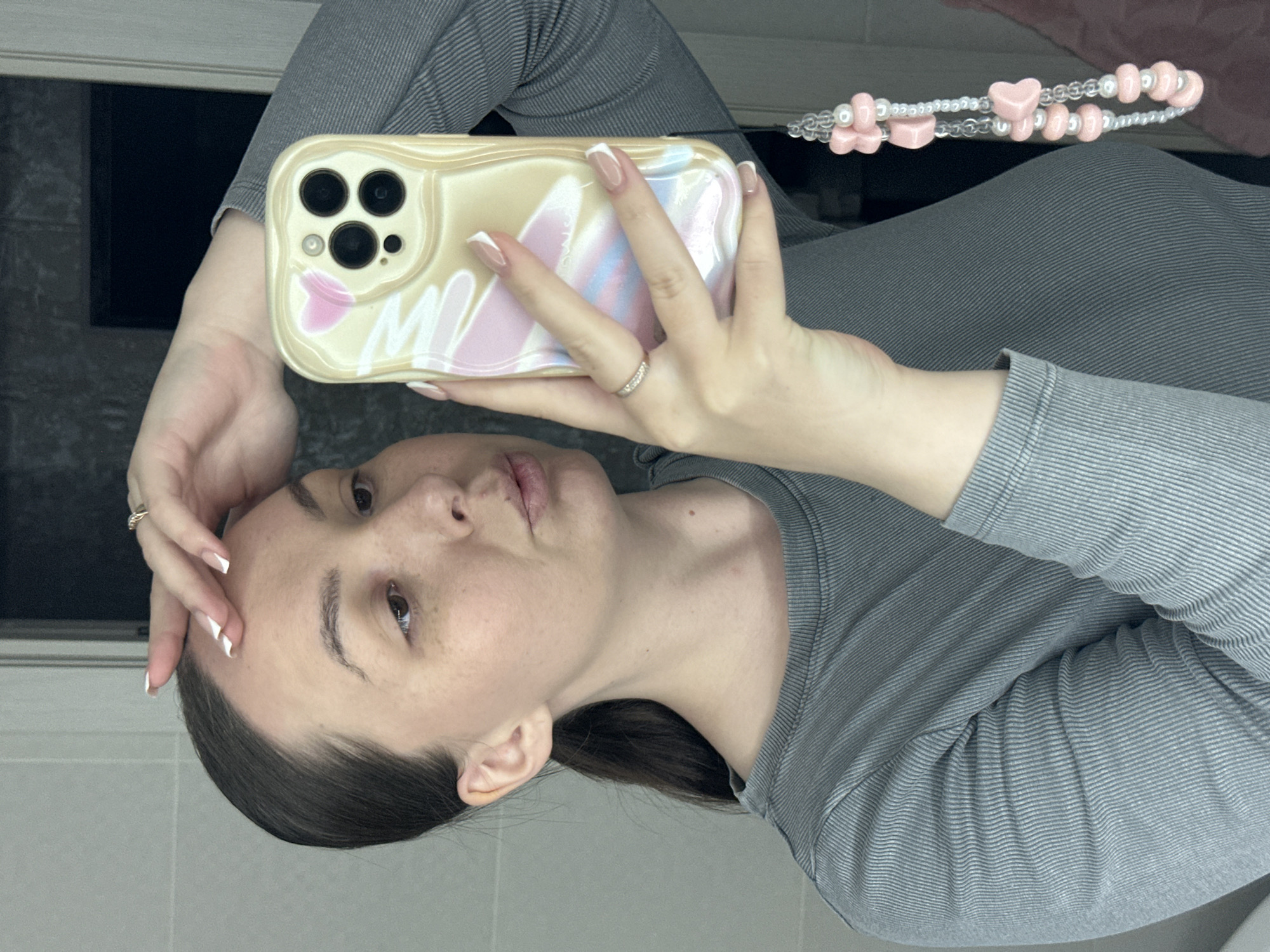 A woman is holding a cell phone up to take a selfie. She has brown hair and fair skin. She is wearing a gray shirt. The cell phone has a light-colored case with a design on it, and a beaded charm hanging from it. She is in what appears to be a bathroom, with light-colored walls. The location is unknown.
