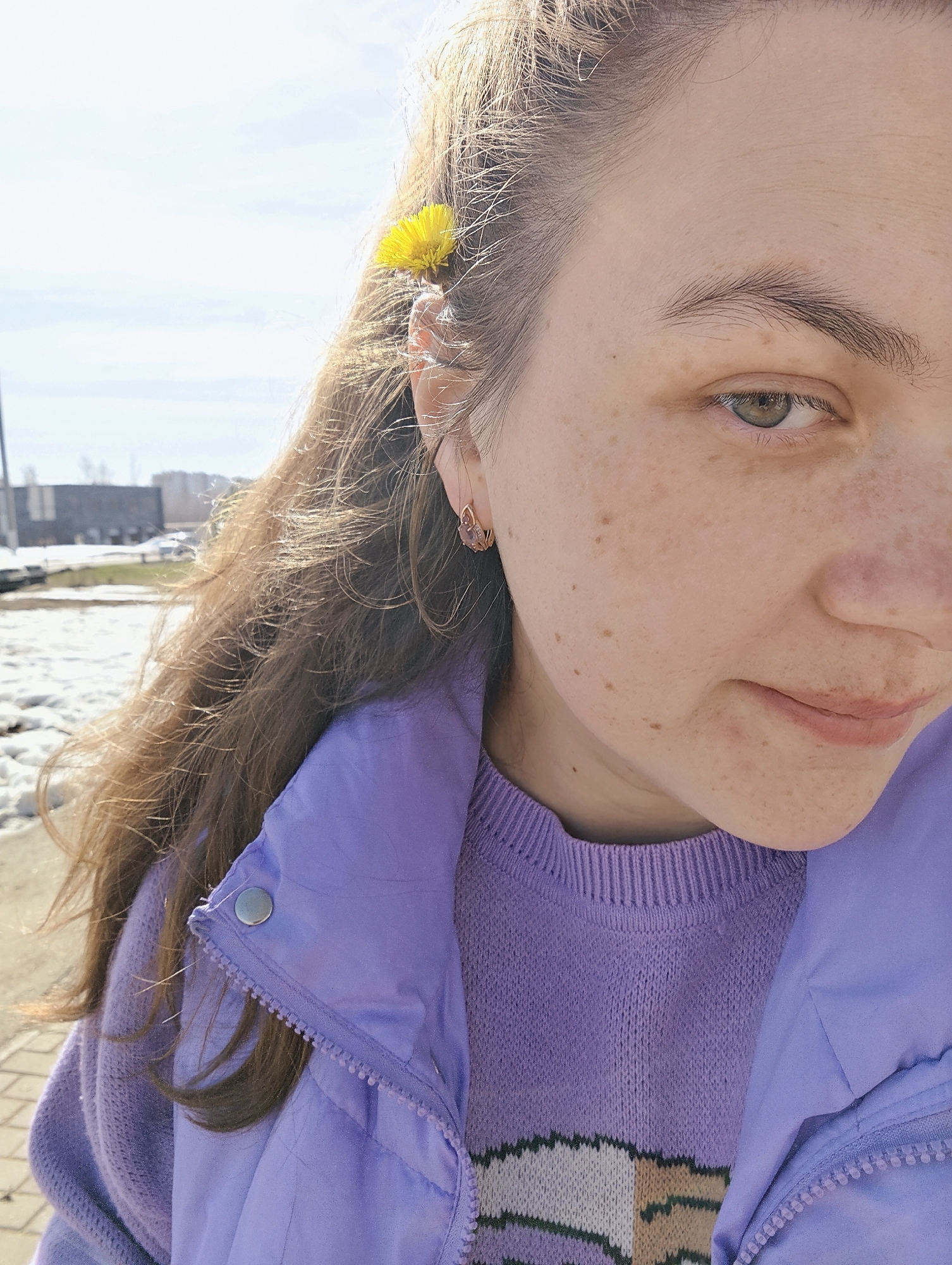 A young woman with fair skin and freckles is pictured in a medium close-up shot. She has brown hair with a yellow flower tucked behind her left ear. She is wearing a light purple sweater and a matching puffer vest. Her left ear is adorned with a small, delicate earring. The woman is looking down and slightly to the right, with a gentle expression on her face.
The background reveals an urban environment. A building is visible, and remnants of snow can be seen on the ground, suggesting it is early spring. The presence of snow and the architecture suggest this location is in Chaikovsky, Russia.