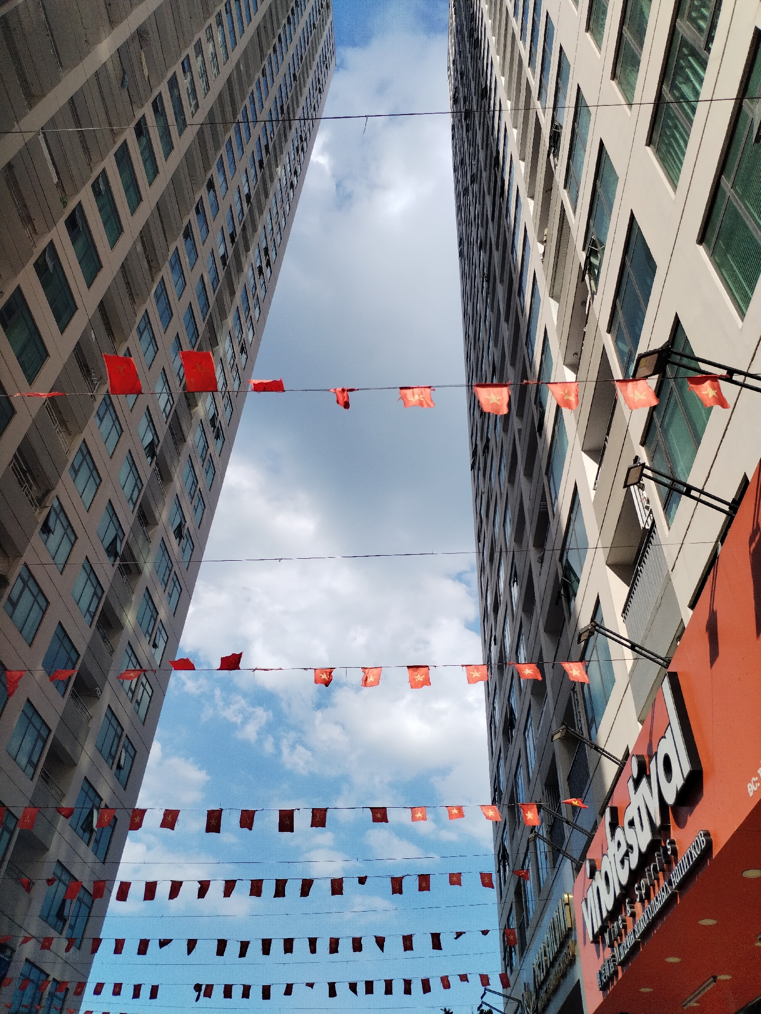 Captured from a low-angle perspective, the image reveals a view of two tall buildings in Bac Nha Trang Ward, Vietnam. A narrow band of sky with white clouds is visible between the buildings. Several lines of small red flags with a yellow star are strung between the buildings. On the right, a commercial sign with the word "Vinotfestival" is partially visible.