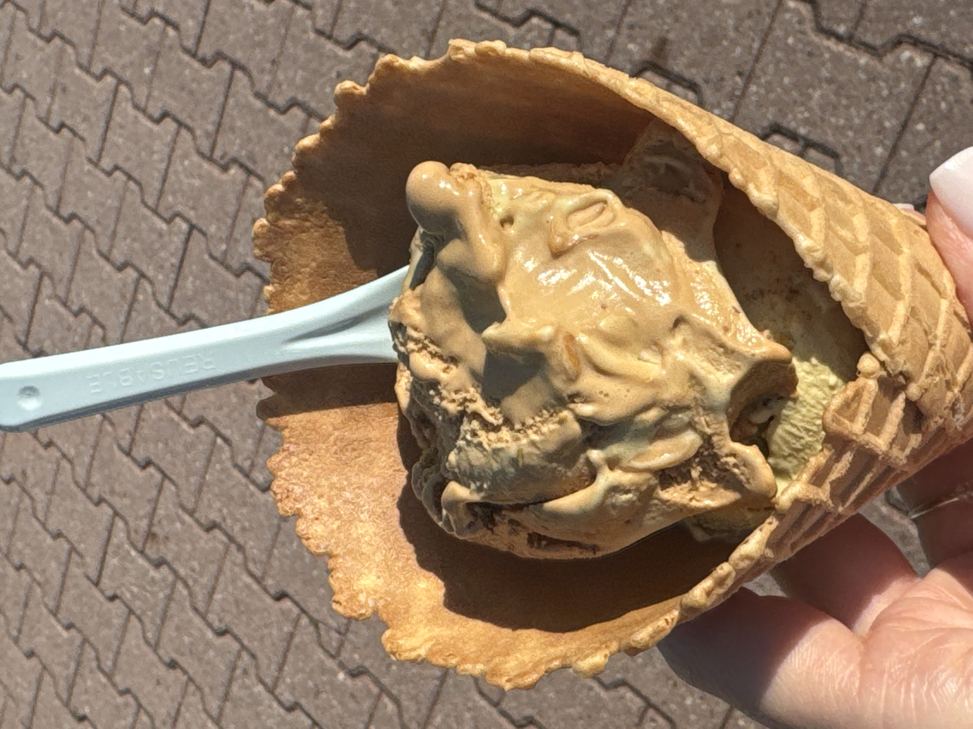 An overhead shot shows an ice cream cone held in a gloved hand. The cone is a waffle cone and is filled with two scoops of ice cream. The top scoop is a caramel or peanut butter flavor, and it is partially melted. The bottom scoop appears to be a lighter green color, possibly pistachio or mint. A light blue plastic spoon is stuck into the ice cream. The background is a brick-like paved surface. There is no indication of the city of Bratislava, Slovakia in the image.
