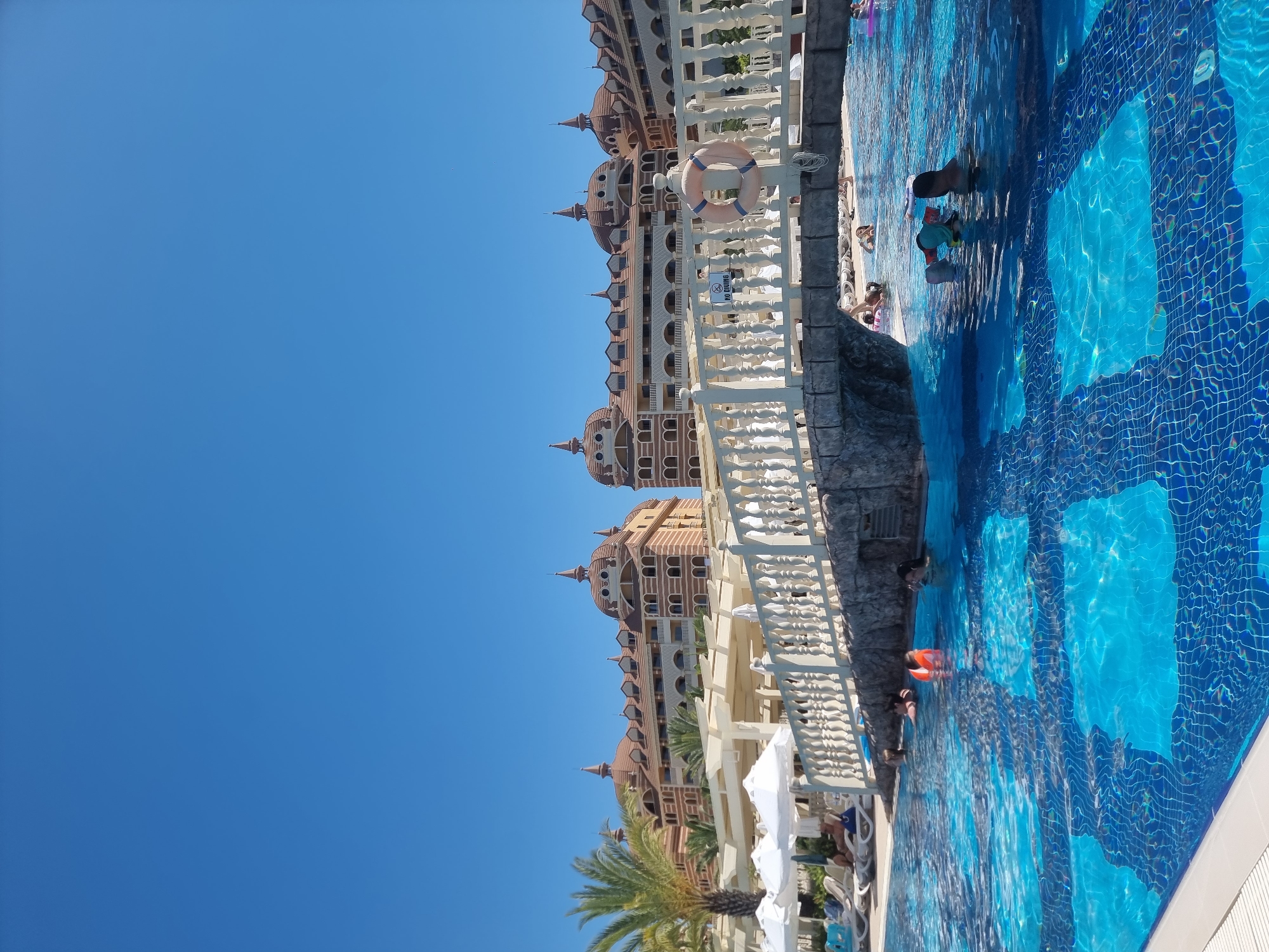 A wide-angle shot captures a resort swimming pool on a sunny day, with a clear blue sky dominating the upper portion of the image. In the foreground, the crystal-clear blue water of the swimming pool is visible, with intricate blue and white mosaic patterns on the pool floor. Several people are enjoying the water; some are partially submerged, while others are playing or relaxing. One person wears an orange life vest. A white bridge with ornate balustrades spans across the pool, with a life preserver hanging on one of its posts. A sign indicating "NO DIVING" is also visible on the bridge. In the background, a large, multi-story resort building with distinctive dome-shaped roofs and intricate architectural details stands against the sky. Palm trees and white umbrellas add to the tropical ambiance of the scene. This appears to be the Aqua Fantasy Aquapark Hotel & Spa in Manavgat, Turkey.