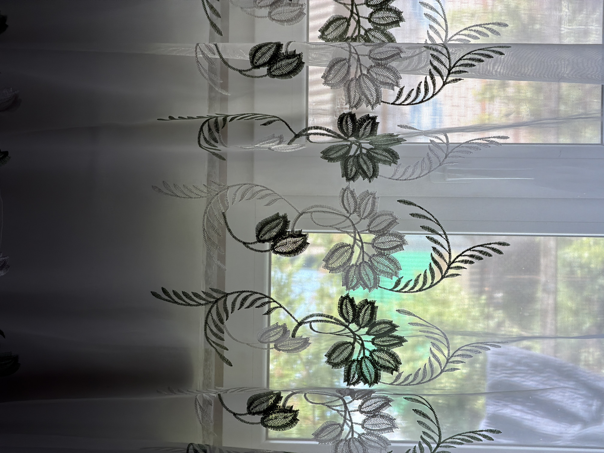 The image captures a close-up view of translucent white curtains covering a window, with a blurred outdoor scene visible beyond. The curtains are adorned with an intricate embroidered pattern featuring stylized foliage and floral motifs in varying shades of grey and dark grey, almost black, creating a textured appearance. The embroidery is dense, giving the leaves and petals a distinct, ridged look.

The curtains themselves appear to have horizontal bands or pleats, allowing light to diffuse through them. Strong daylight streams in from the window, illuminating the sheer fabric and highlighting the embroidered designs against the bright background. The outdoor view, though out of focus, shows vibrant green foliage, suggesting a garden or natural setting. Hints of brighter colors, possibly blues or lighter greens, are also discernible in the blurred background. The left side of the image is significantly darker than the right, indicating a shadow or less direct light reaching that portion of the curtain. No people, animals, or visible text are present in the frame. The scene conveys a sense of peaceful daytime and a connection between an indoor space and the natural environment.