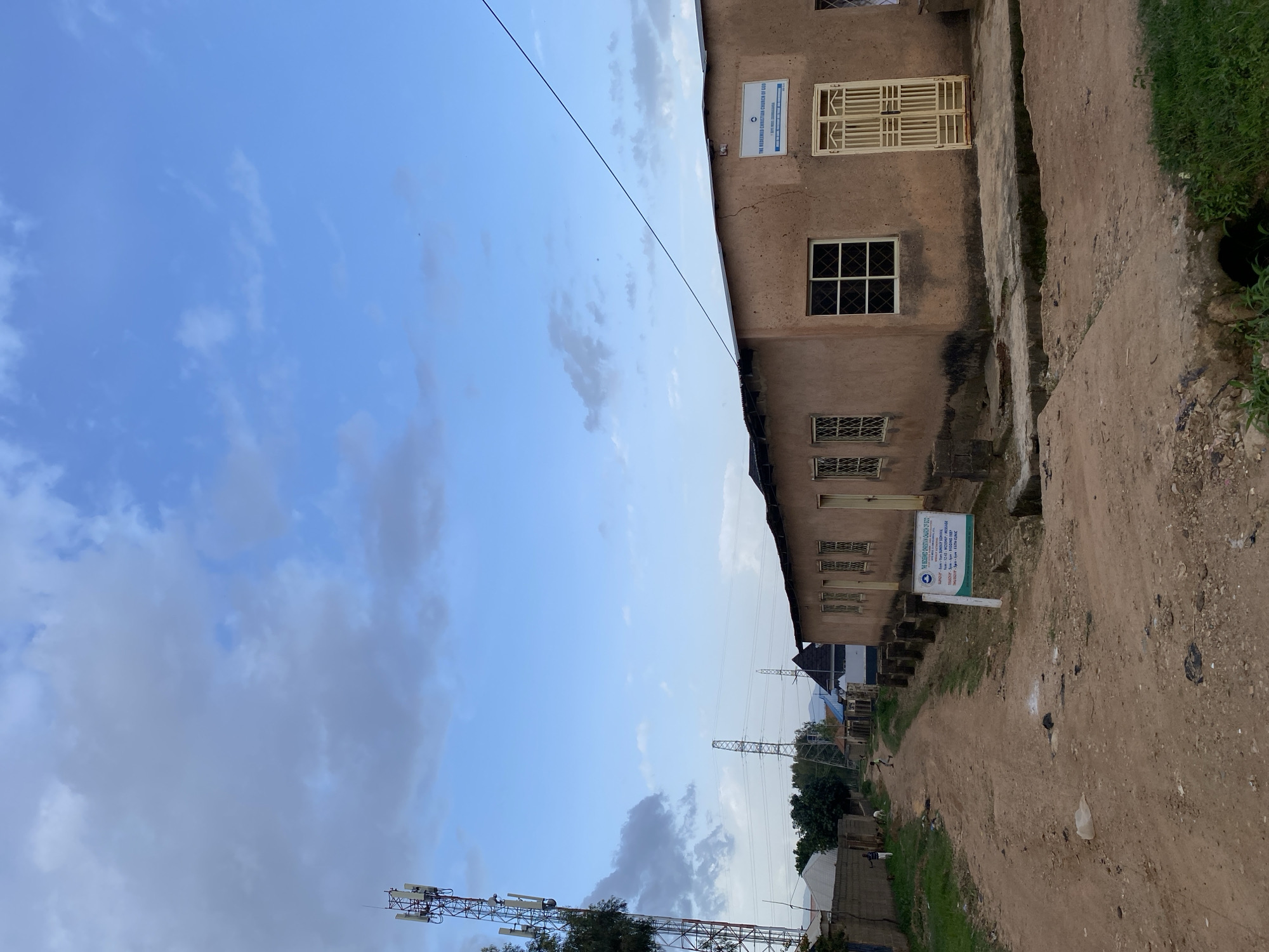 A dirt road in Gura Top, Nigeria, leads past a building identified as "THE REDEEMED CHRISTIAN CHURCH OF GOD." The building appears to be constructed with mud and has a corrugated metal roof. It features several windows with decorative metal grates and a yellow metal door. A sign on the front of the building lists service times for Sunday and Tuesday, and mentions "FAITH CLINIC" on Thursday. In the background, a tall cellular tower stands beside a street. The sky above is partly cloudy with a blue hue. Two children are visible in the distance playing on the dirt road.