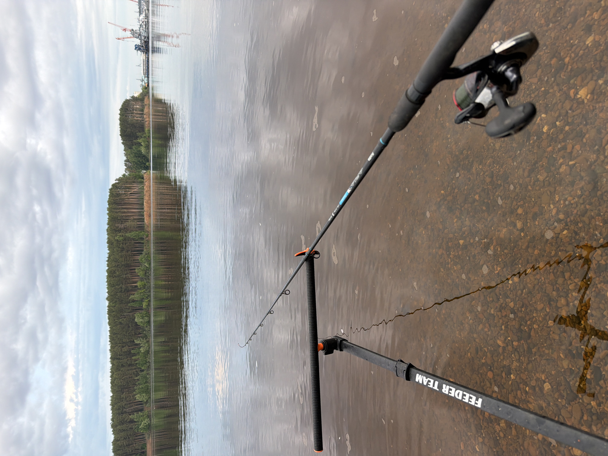The image shows a fishing rod set up on the bank of a body of water in Chaikovsky, Russia. The rod is black with an orange support and a reel attached. The water is murky, with the bottom visible and what appears to be a reflection. The background includes a treeline with a hint of industrial infrastructure on the horizon. The sky is overcast. The fishing line extends into the water. The side of the fishing rod has "FEEDER TEAM" written on it.