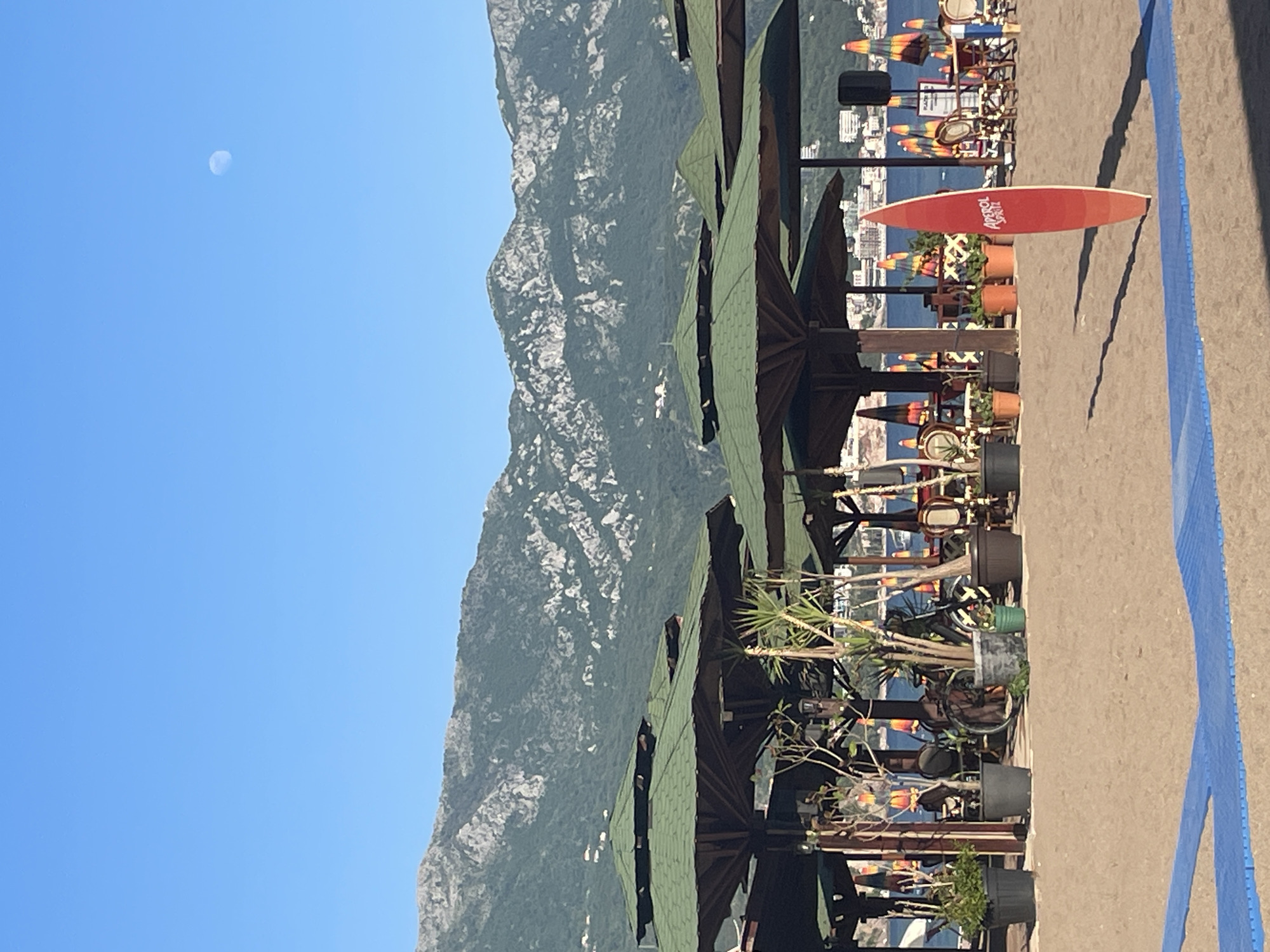 The image depicts an outdoor scene in Boreti, Montenegro, against a backdrop of mountains under a clear blue sky with the moon visible. Foreground elements include a series of parasols, potted plants, tables and chairs, suggesting a beachfront cafe or lounge. In the distance, colorful beach umbrellas and deck chairs mark the shoreline, with a structure or building visible beyond. A red paddleboard with the word “Aperol” on it lies on the sandy ground. A blue boundary line is also visible.