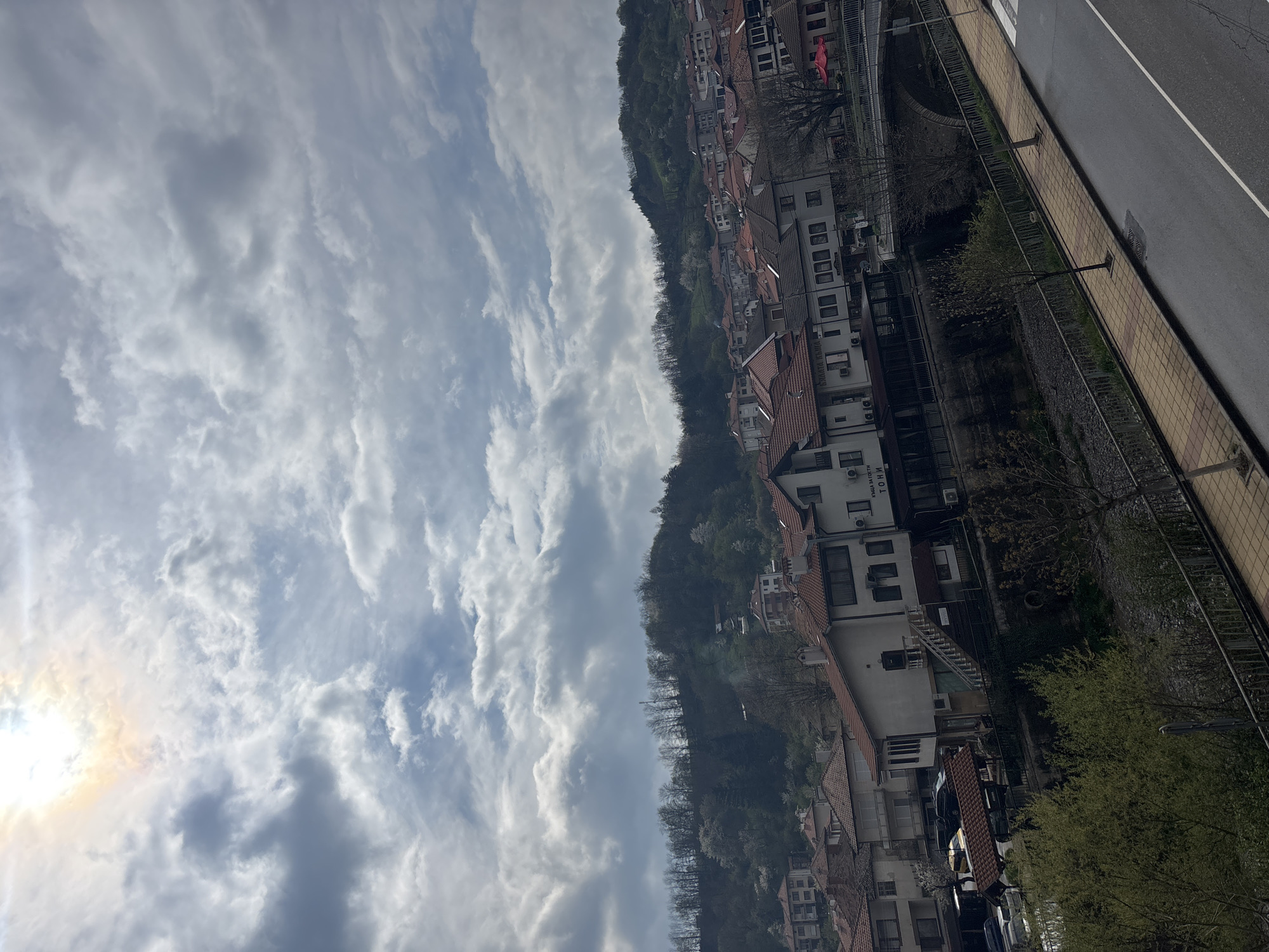 The image presents a scenic view of a town nestled on a verdant hillside under a dynamic, partly cloudy sky. The setting, identified as Zlatograd, Bulgaria, features numerous buildings with traditional red-tiled roofs, scattered across the green, tree-covered slopes. Many structures have white or light-colored facades, with some displaying modern amenities like air conditioning units. The hillside is densely forested with a mix of trees, some appearing bare while others are green, suggesting early spring or late autumn.
The sky fills a substantial portion of the upper frame, dominated by an array of white and grey stratocumulus clouds. A bright sun, partially obscured by clouds on the left, creates a strong glare and shows a faint iridescence, indicating it is daytime, likely mid-morning or afternoon.
In the foreground, on the right, a paved road with a white demarcation line runs alongside a patterned tiled sidewalk. A metal fence separates the sidewalk from a lower, greener area that features more trees and additional building roofs. A street lamp is visible on the sidewalk. No people or specific activities are taking place, portraying a calm, static landscape. Visible Cyrillic text on a building in the lower-middle reads "КЪЩА ЗА ГОСТИ ТОНИ" (Guest House Toni).