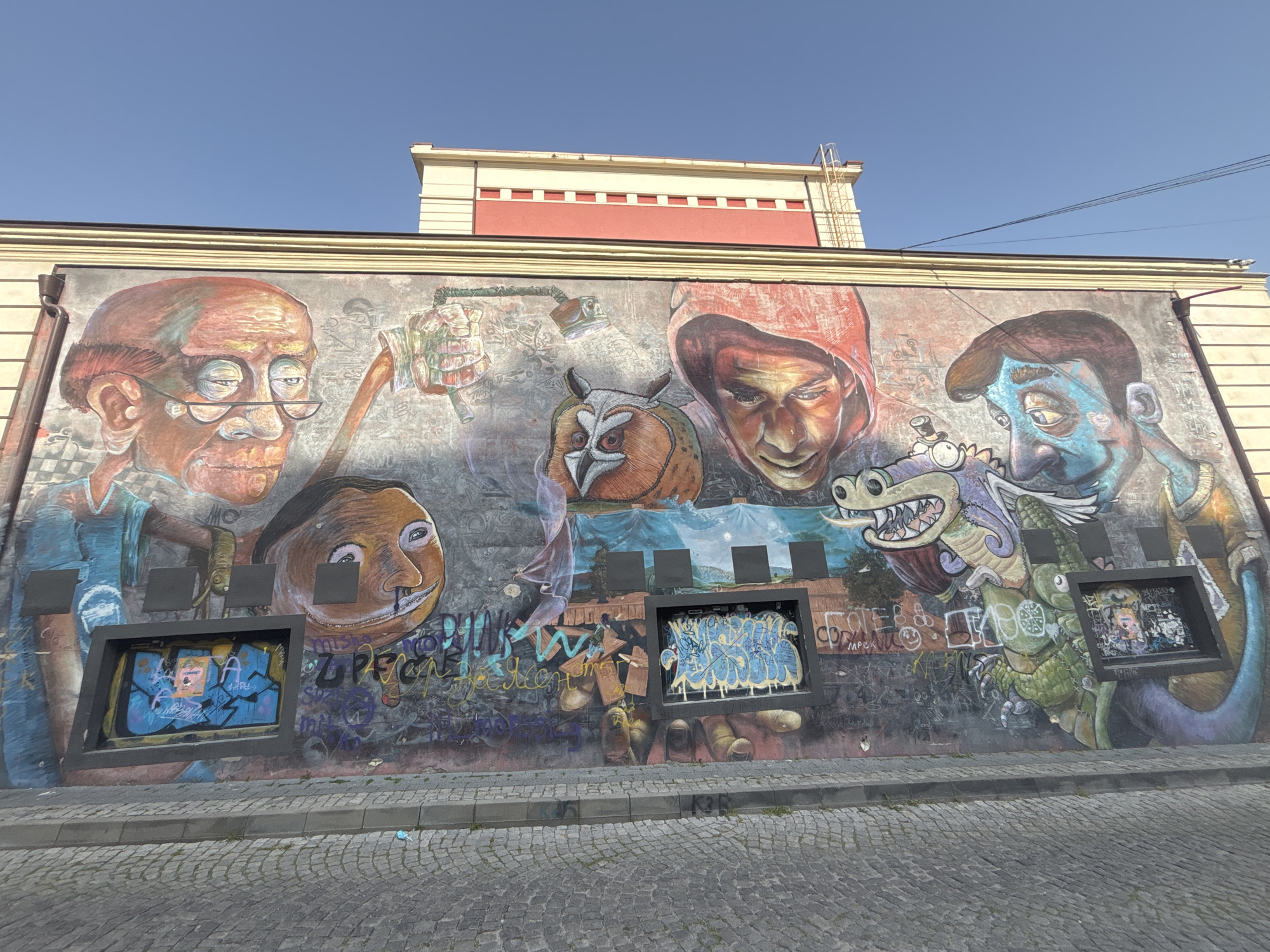 The image captures a mural in Plovdiv, Bulgaria, featuring a variety of painted figures. From left to right, the mural includes a portrait of an older man wearing glasses, a smiling face, an owl, a person wearing a red hood, and a blue-skinned individual alongside a purple and green dragon. The artwork is interspersed with graffiti. Embedded within the mural are three dark, square frames, each containing distinct painted designs. The mural is painted on the side of a building. A cobblestone road runs along the front of the mural. A cream and red building is visible above the mural.