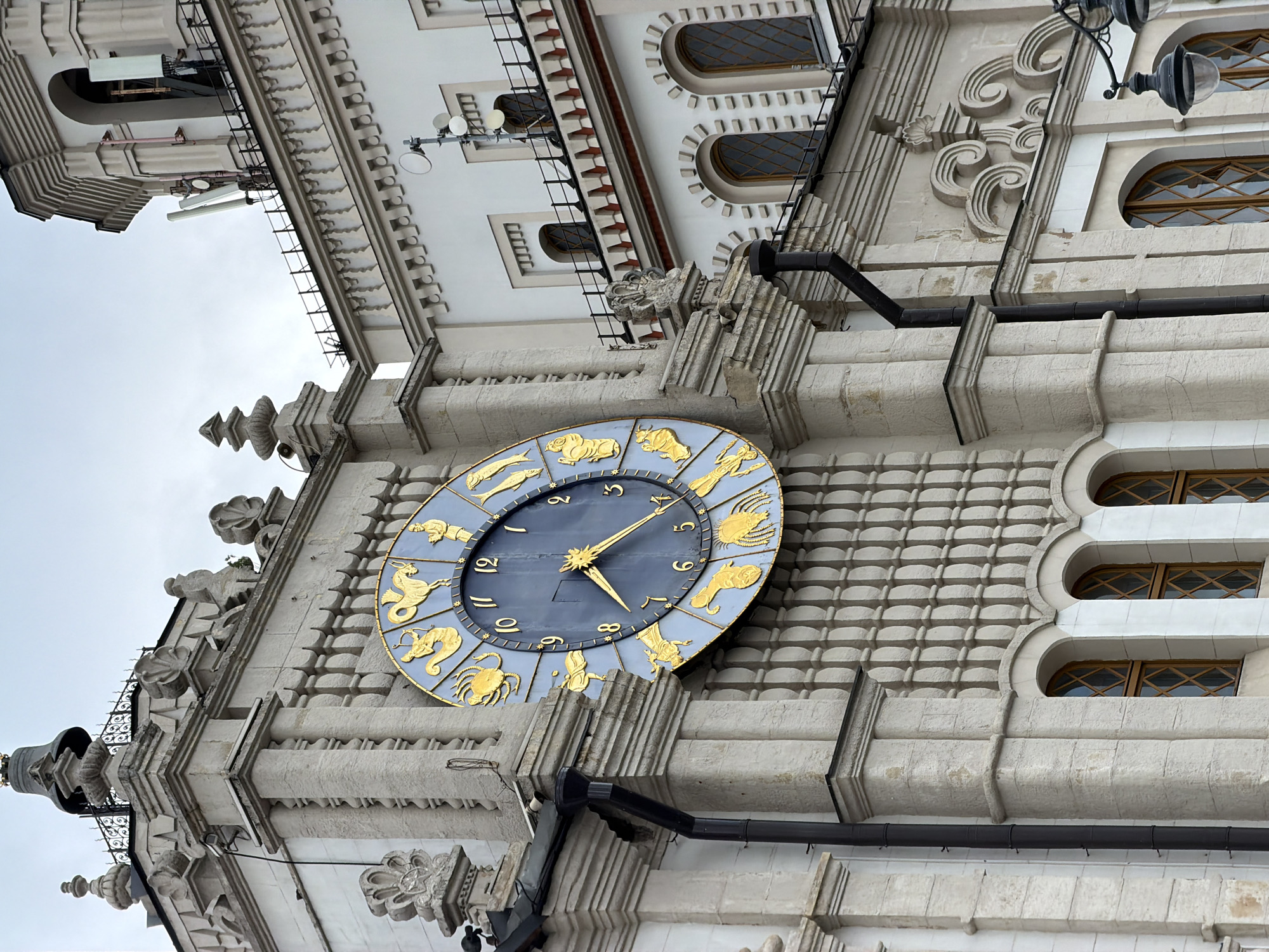 A low-angle shot shows the clock on the building of the State Academic Central Puppet Theatre named after S.V. Obraztsov in Moscow, Russia. The clock, circular with a blue face, is decorated with gold-colored zodiac symbols around the edge. The building's facade, rendered in light-colored stone, features intricate architectural details, including carved moldings, arched windows, and decorative elements. A section of a black drainpipe runs along the building's exterior near the clock. The sky appears overcast in the background.