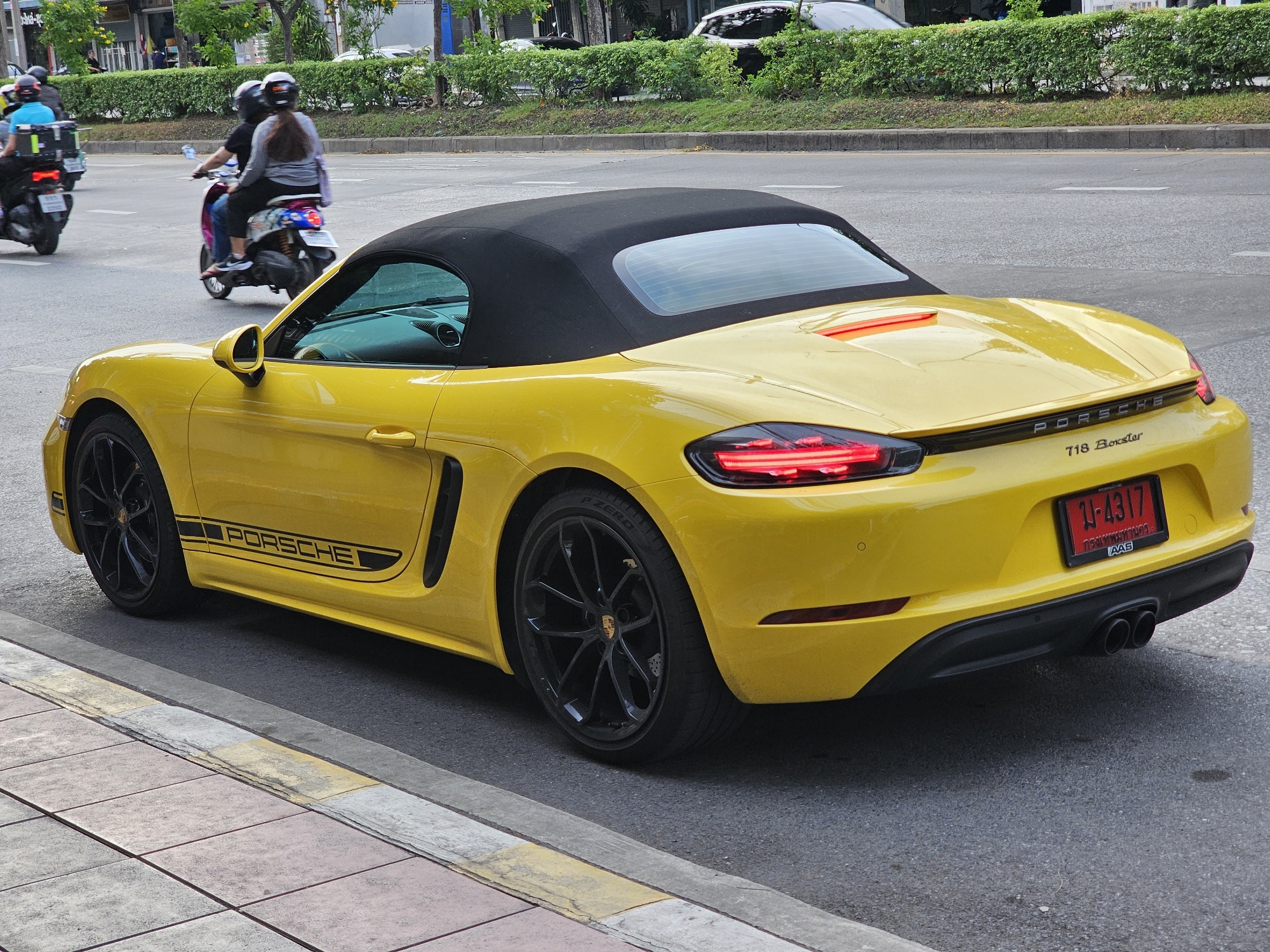 A bright yellow Porsche 718 Boxster convertible is parked on the right side of a street in Bangkok, Thailand. The car has a black soft top and black rims on its wheels. The license plate is red with white Thai characters and numbers "14317". The car is on a paved road next to a sidewalk with patterned tiles. In the background, there is a traffic island with manicured green bushes and trees lining the road. Several motorcycles are visible in the background, some with passengers wearing helmets. One motorcycle on the far left has a blue-shirted rider and a large black box on the back. Another motorcycle in the middle has two people wearing helmets. The scene appears to be a city street with moderate traffic.