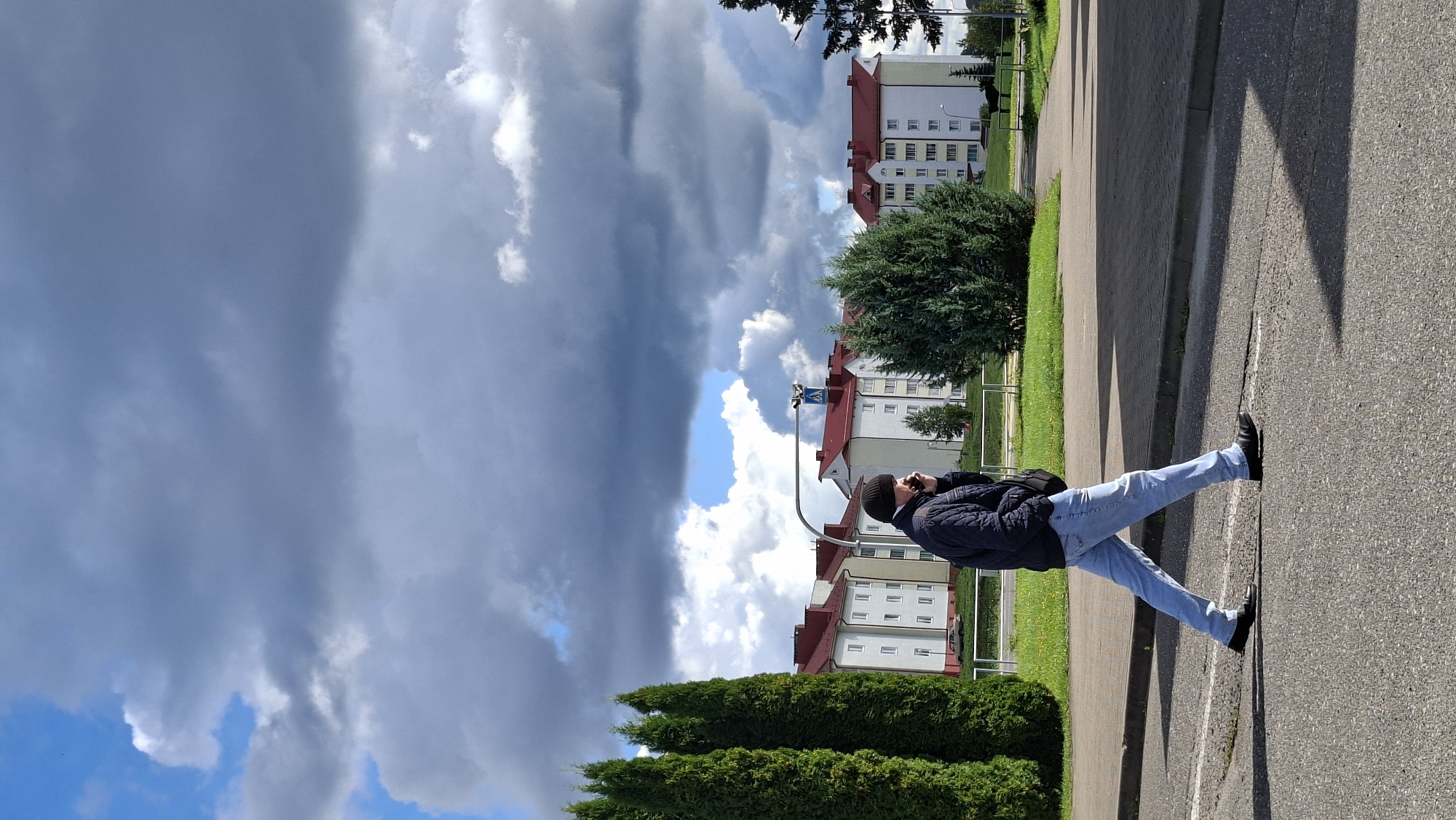 A person stands on an asphalt path next to a grassy area with trees. Buildings with red roofs are visible in the background. The sky is partly cloudy with dark storm clouds and bright blue sky. The person is wearing a dark jacket, light blue jeans, a black beanie, and black shoes. They appear to be holding a phone to their ear. The sun casts a long shadow to the right of the person. The image appears to have been taken during the daytime, possibly late afternoon, given the long shadows. The weather appears to be changing, with dark clouds suggesting an approaching storm or recent rain. The location is identified as Myadzyel, Belarus. A pedestrian crossing sign is visible near the buildings.